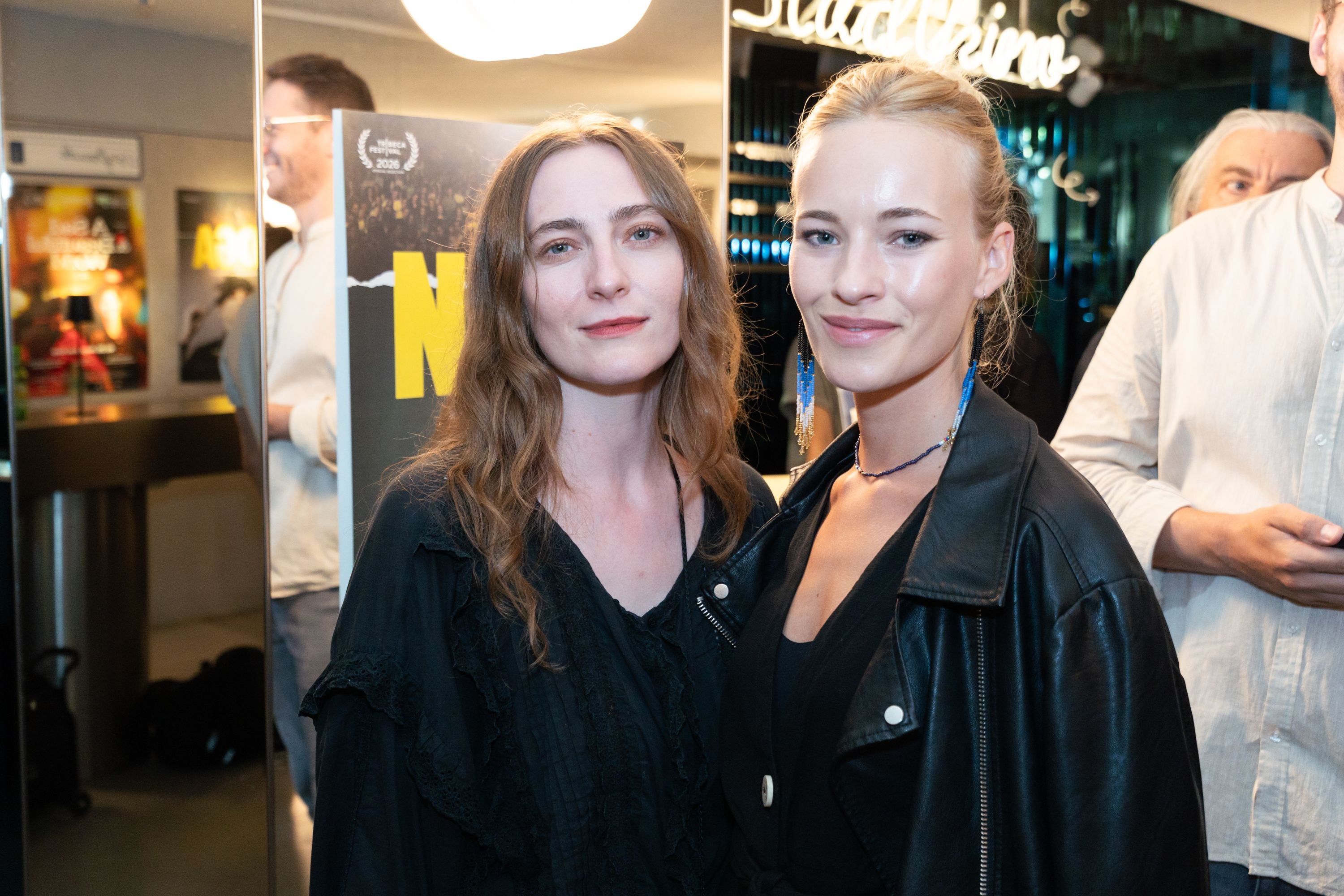 Zwei Frauen posieren bei der NOGA-Premiere im Innenraum vor einem Plakat.