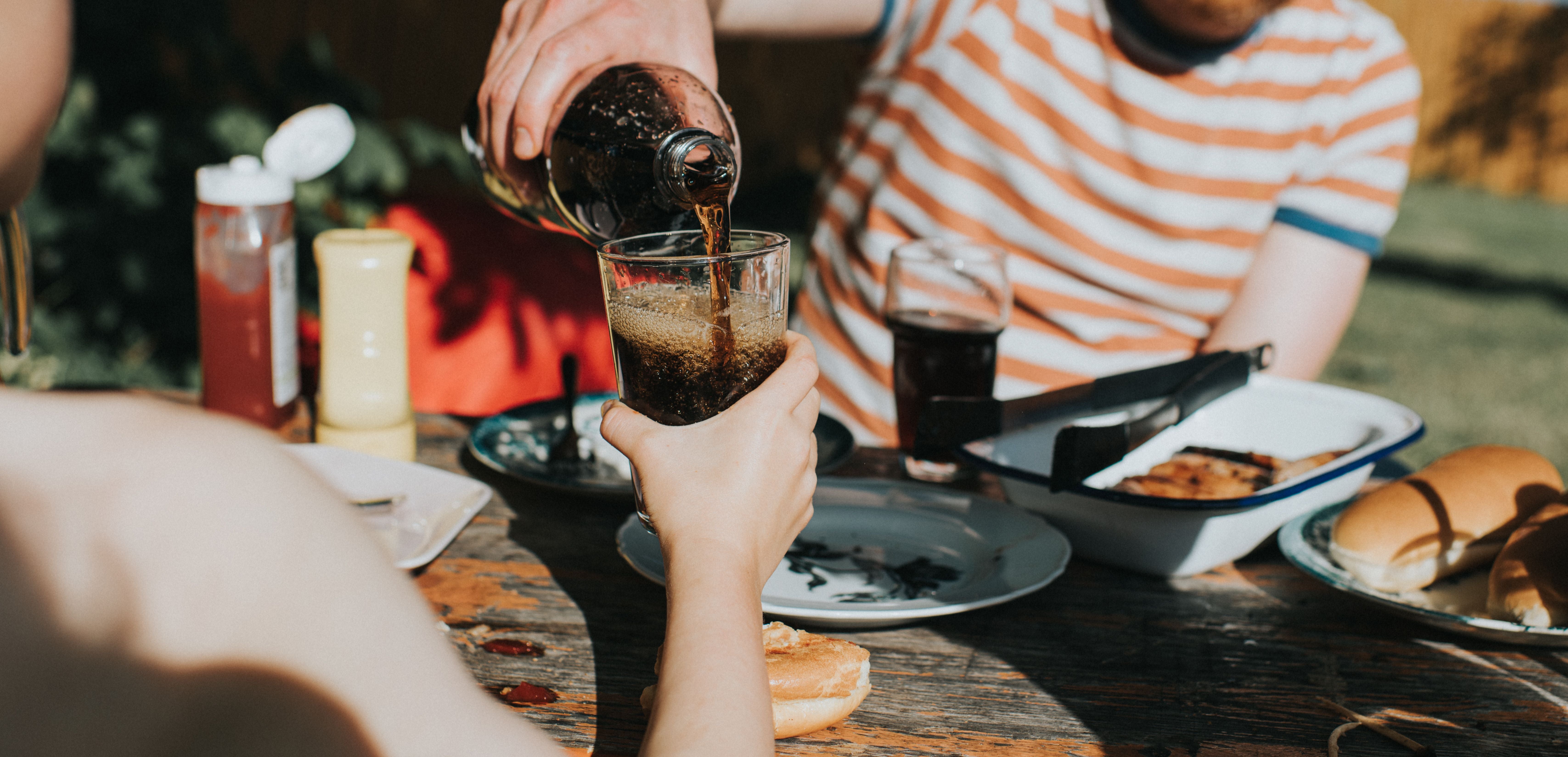 Ein Kind hält ein Glas, das von einem Mann mit Cola an einem Grilltisch draußen gefüllt wird.