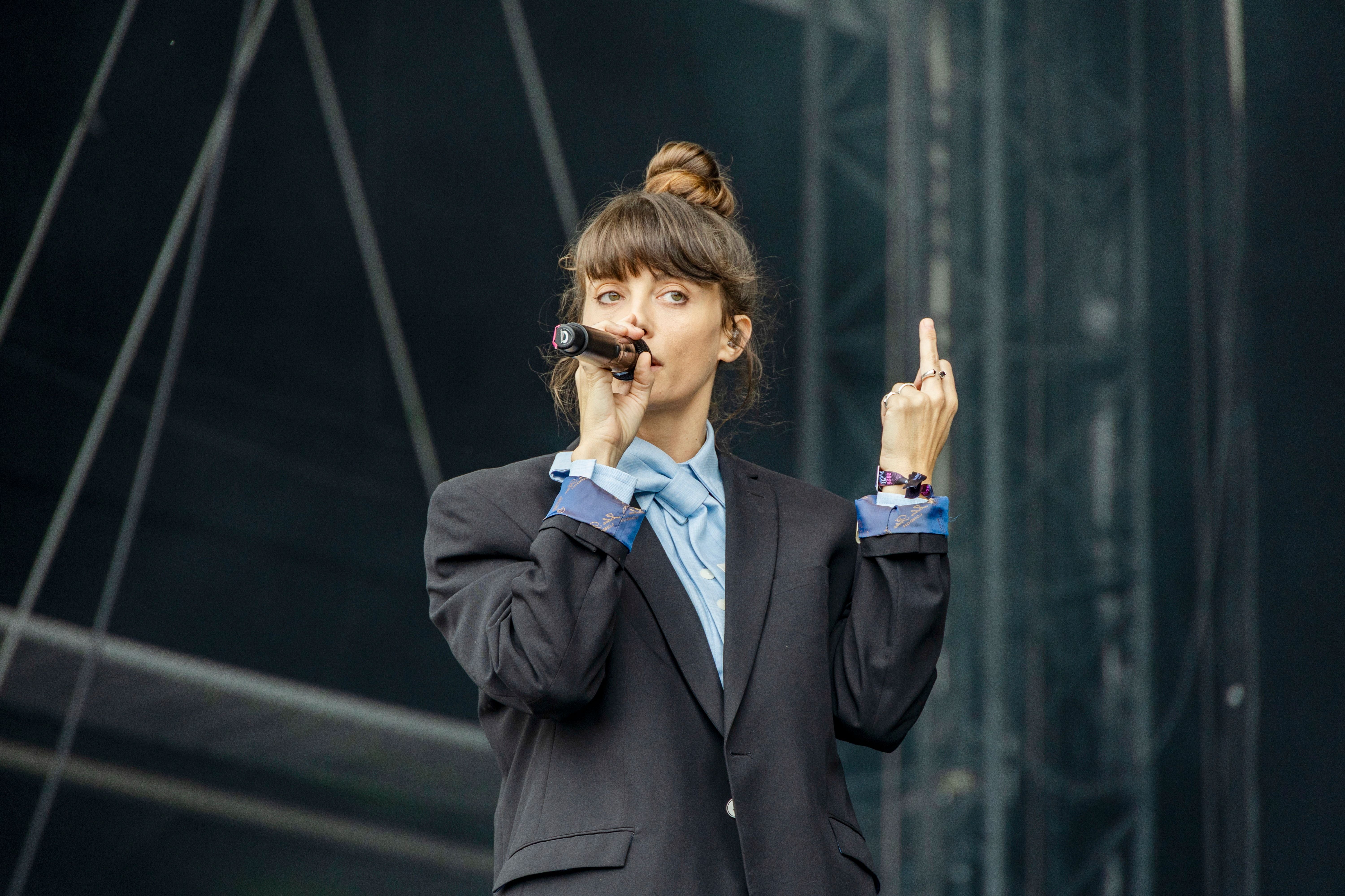 Frau im Anzug singt auf einer Bühne und zeigt den Mittelfinger.