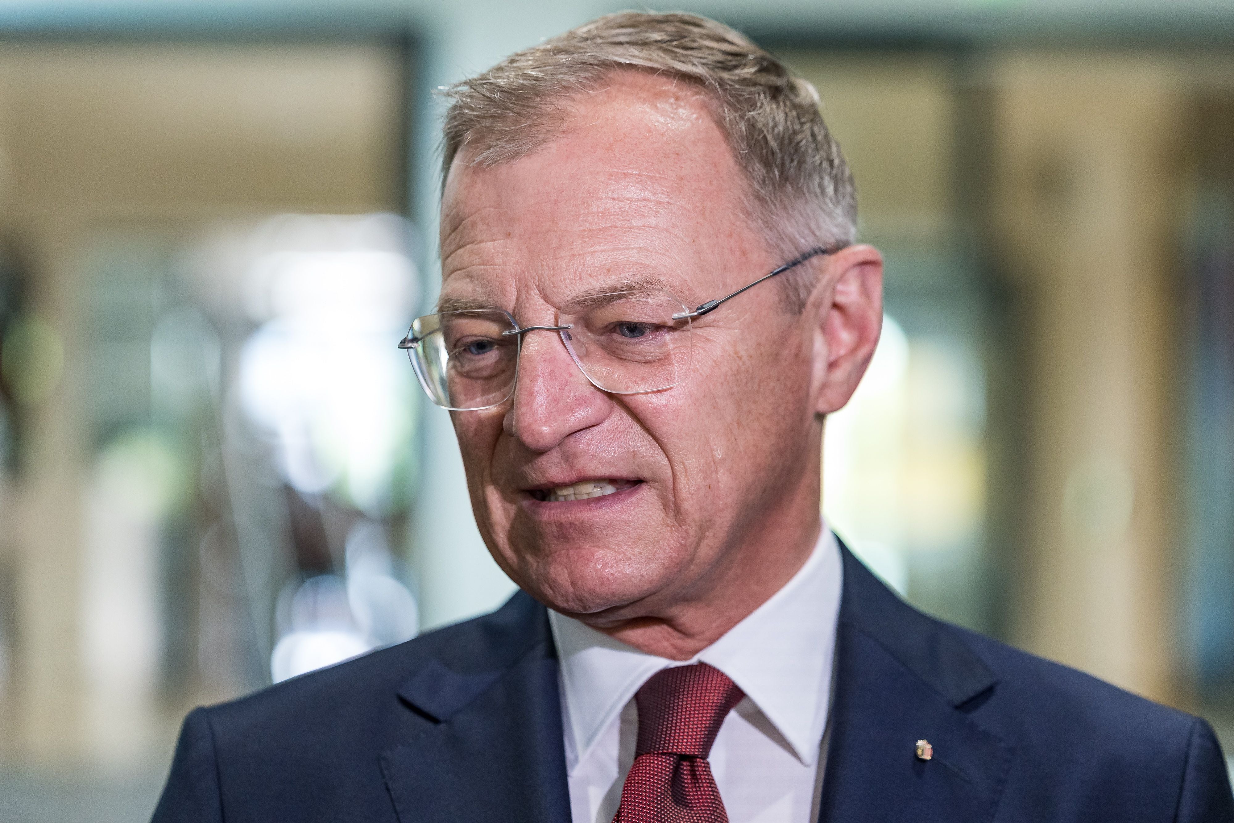 Ein Mann im Anzug mit Brille spricht, unscharfer Hintergrund, Innenaufnahme.