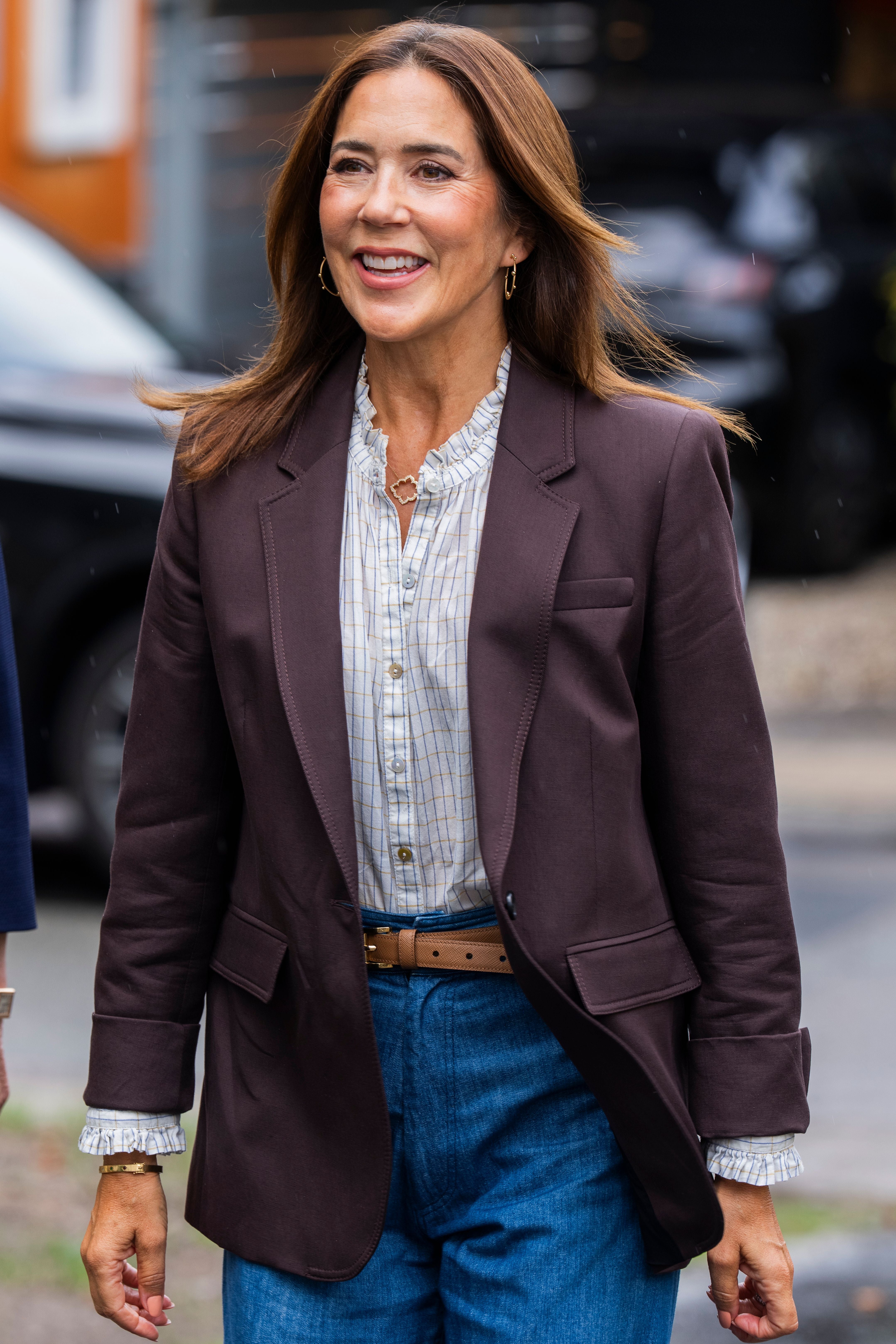 Frau mit braunem Blazer, kariertem Hemd und Jeans lächelt im Freien.