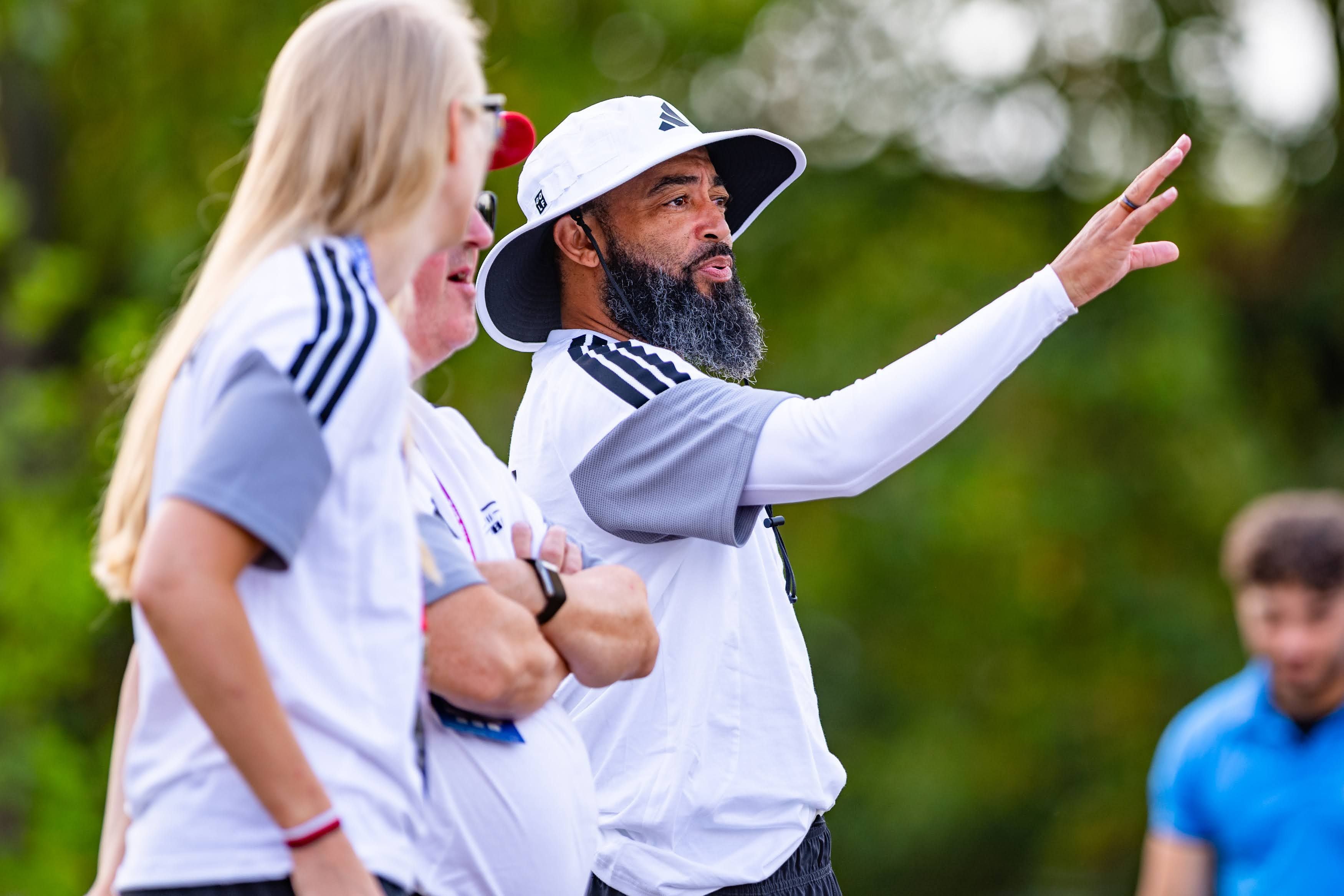 Trainer mit weißem Hut gibt Anweisungen bei einem Flag Football-Spiel im Freien.