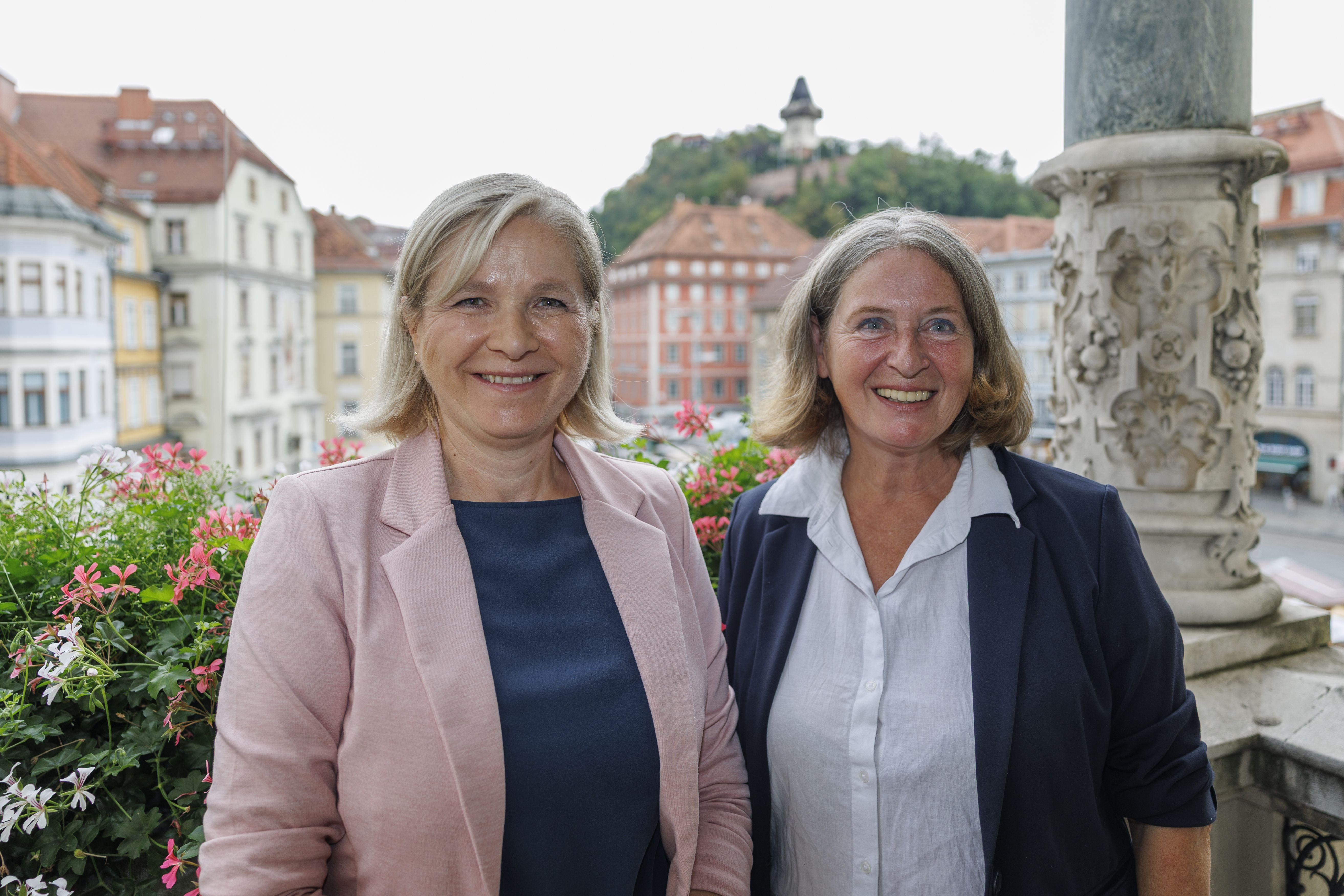 Zwei Frauen lächeln auf einem Balkon mit Blick auf Gebäude und den Grazer Uhrturm.