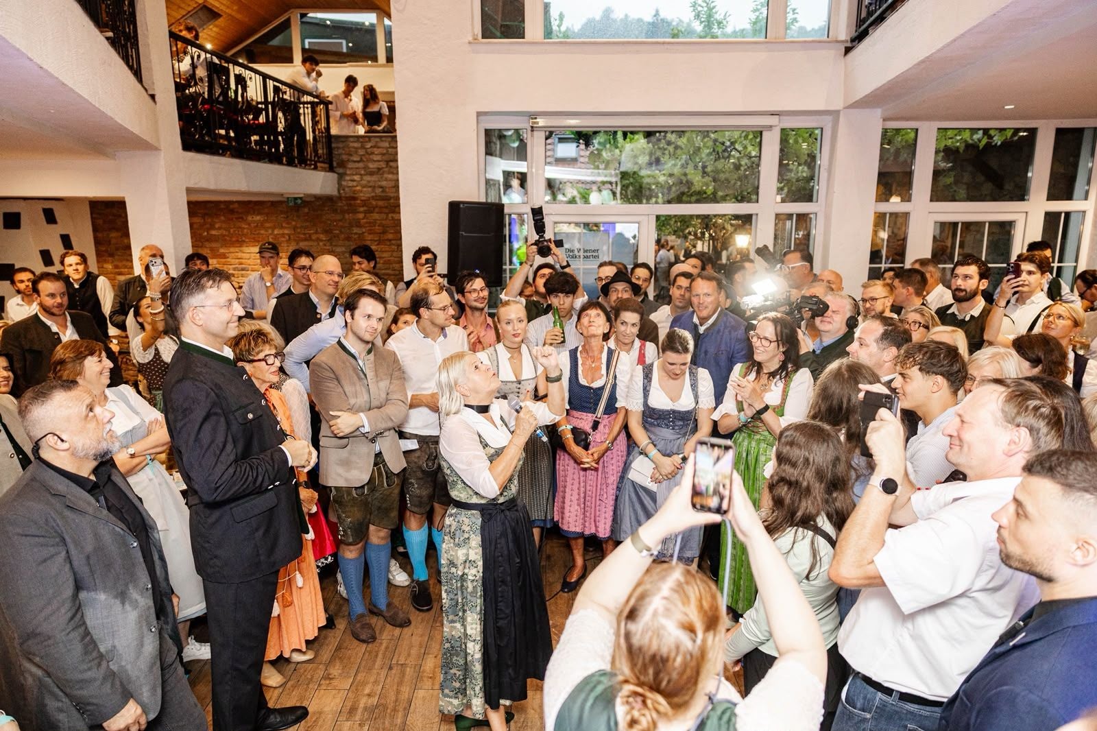 Viele Menschen in Tracht und Anzug stehen bei einer Veranstaltung in einem hellen Saal zusammen.