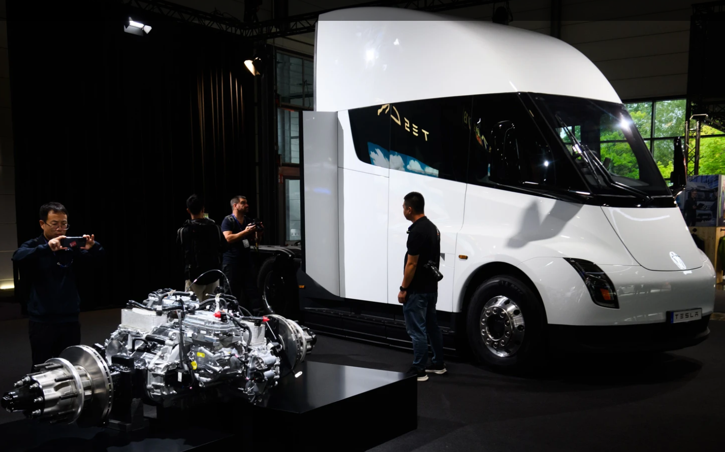 Tesla E-Laster und ein Antriebsstrang werden auf einer Messe von Besuchern betrachtet.