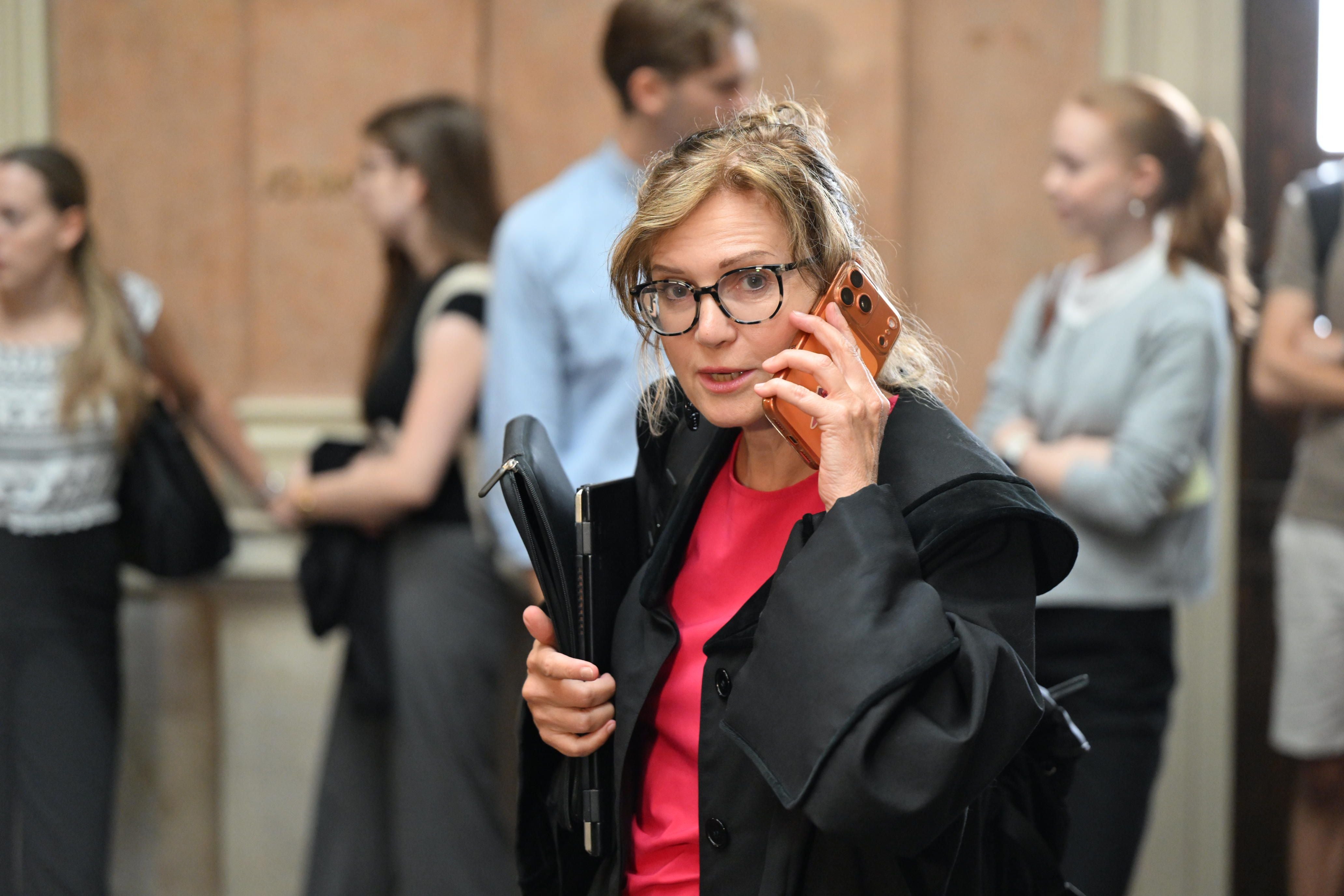 Frau mit Brille telefoniert und hält Dokumentenmappe, im Hintergrund mehrere Personen.
