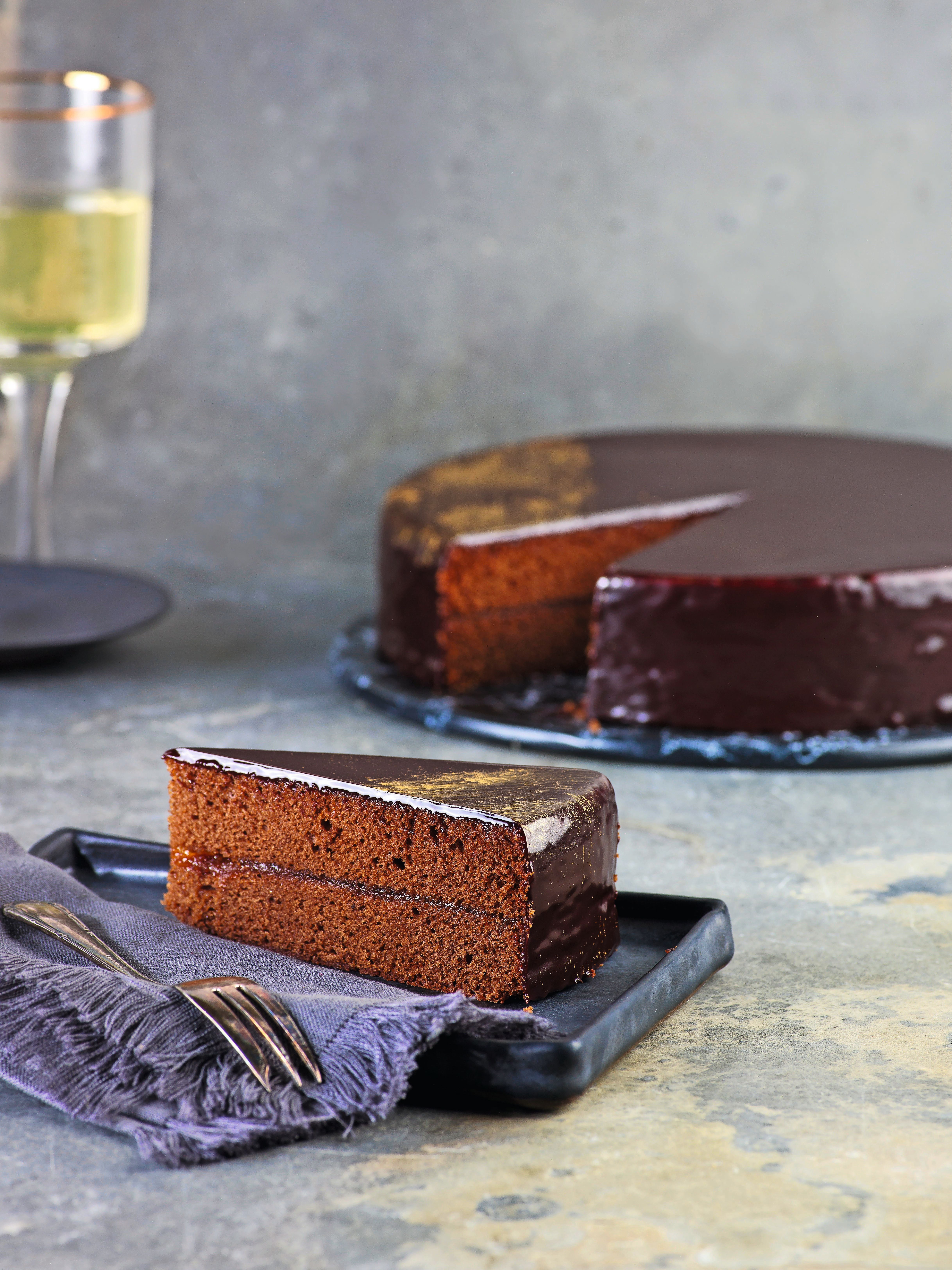 Ein Stück Sachertorte mit glänzender Schokoladenglasur auf einem Teller, daneben Besteck.