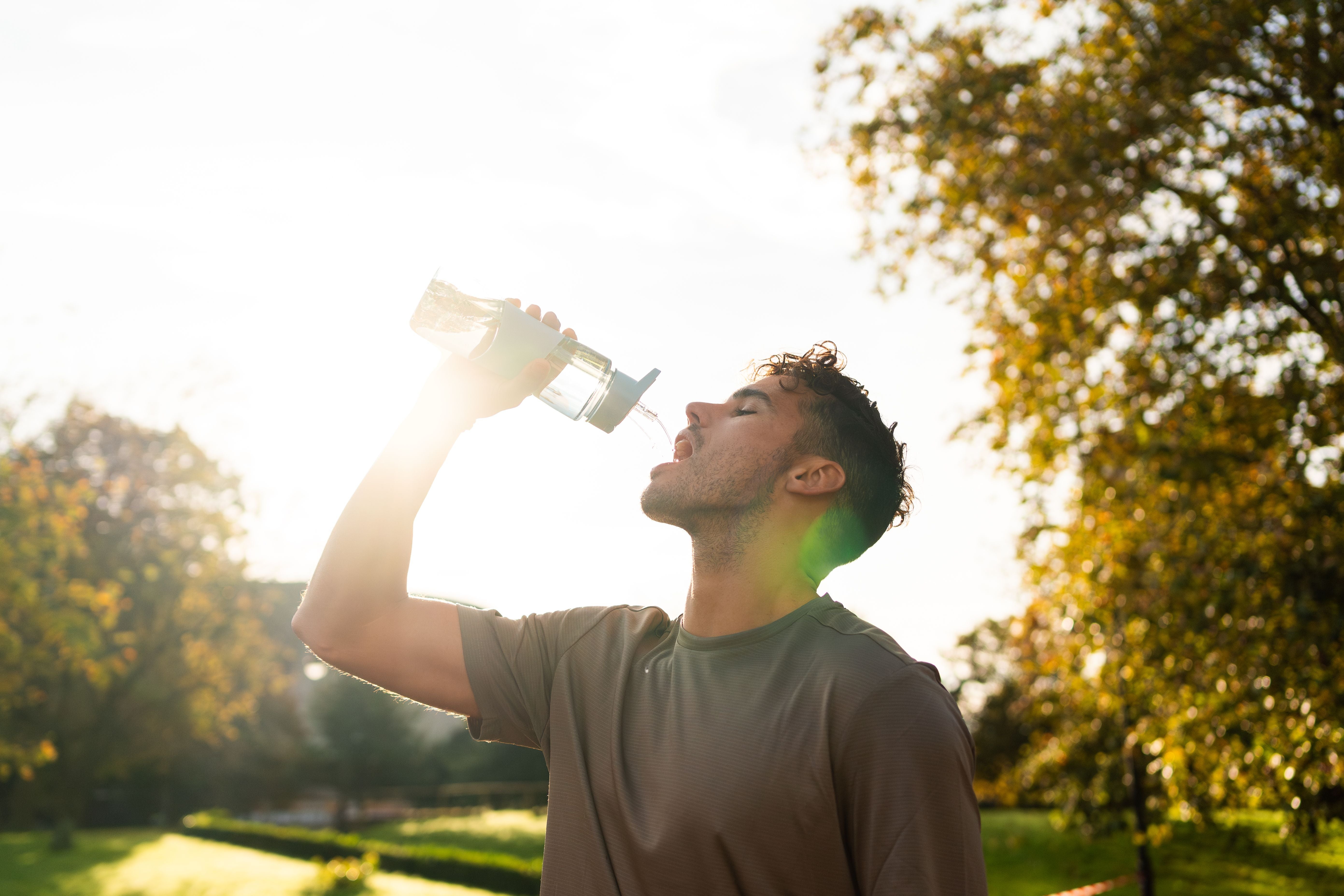 Junger Mann trinkt nach dem Sport im Sommerpark Wasser aus einer Flasche bei Sonnenuntergang.