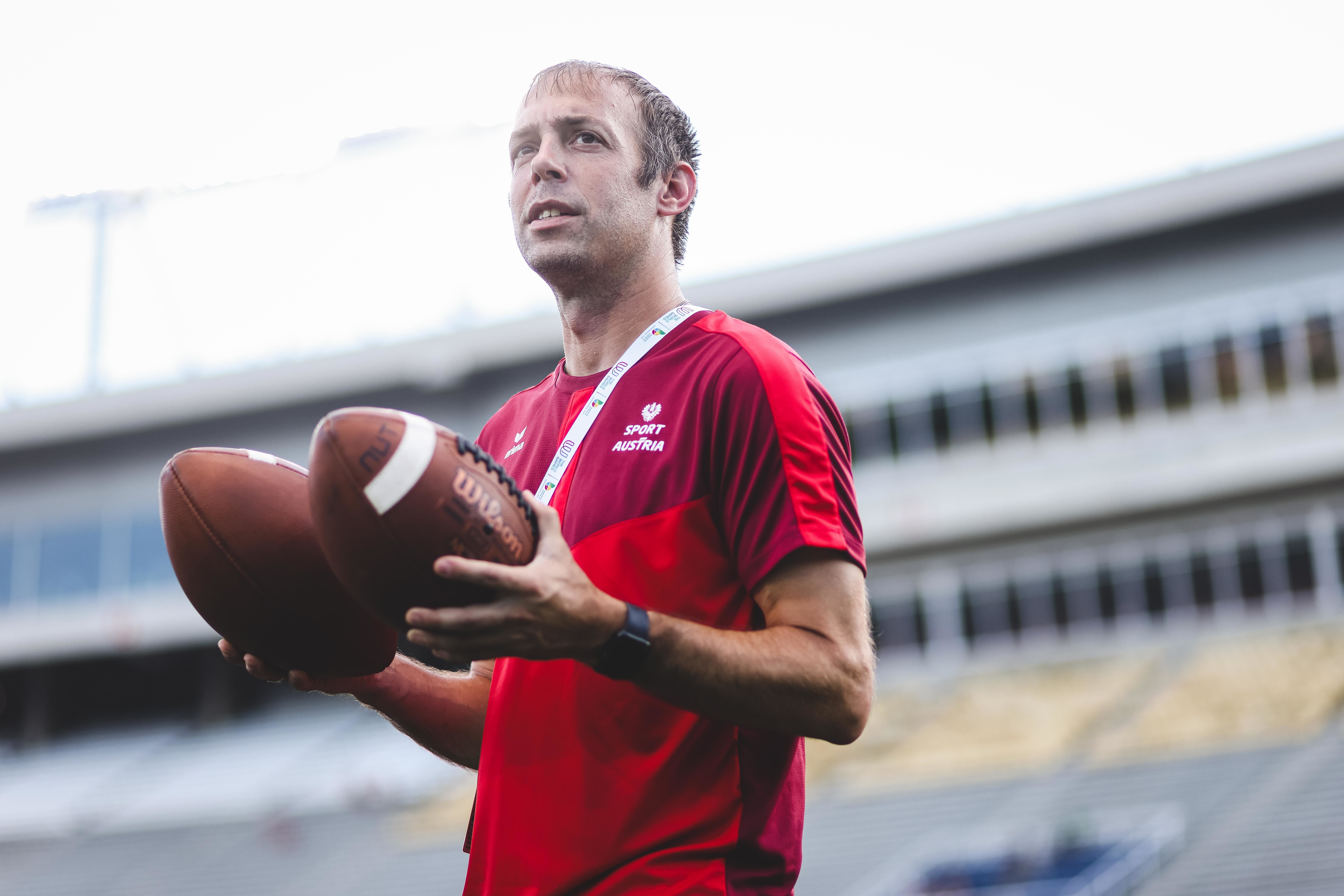 Ein Mann im roten Sportshirt hält zwei American Footballs in einem Stadion.