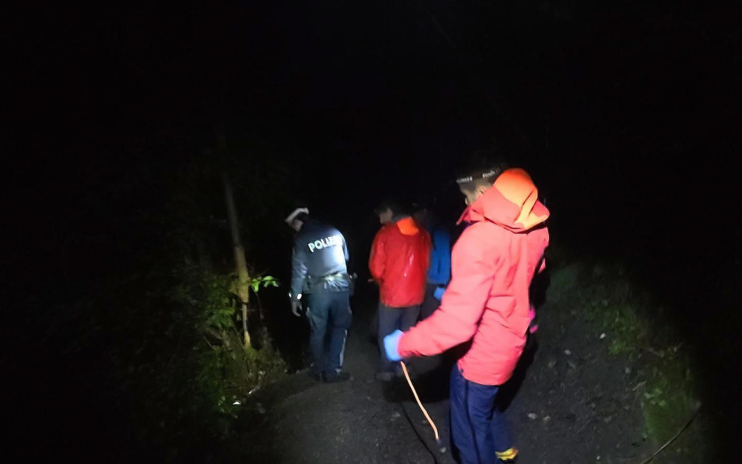 Personen in Outdoor-Kleidung und ein Polizist suchen nachts mit Stirnlampen im Wald.