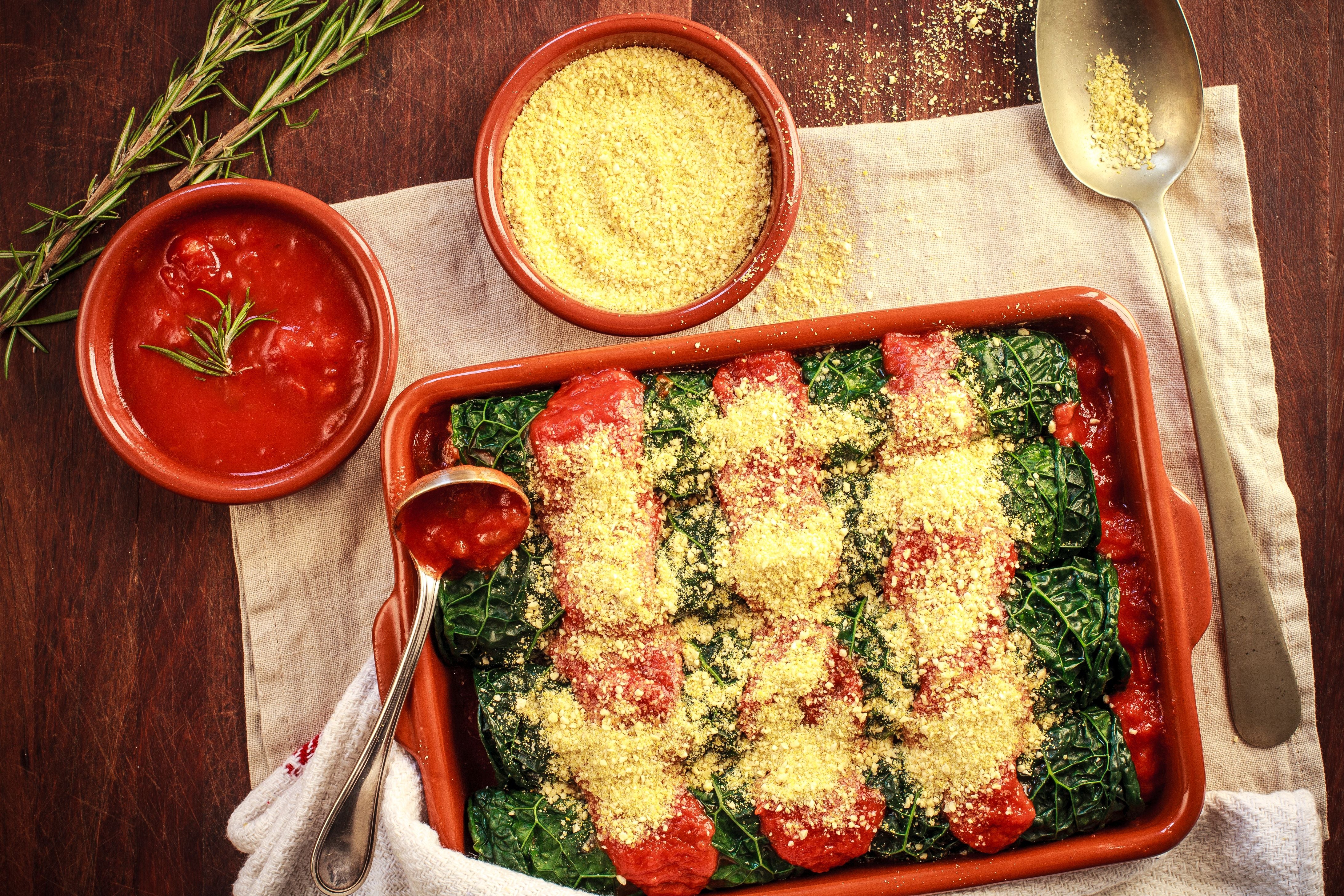 Ein Auflaufform mit Grünkohl-Cannelloni, Tomatensoße und Parmesan, daneben Schüsseln mit Soße und Käse.