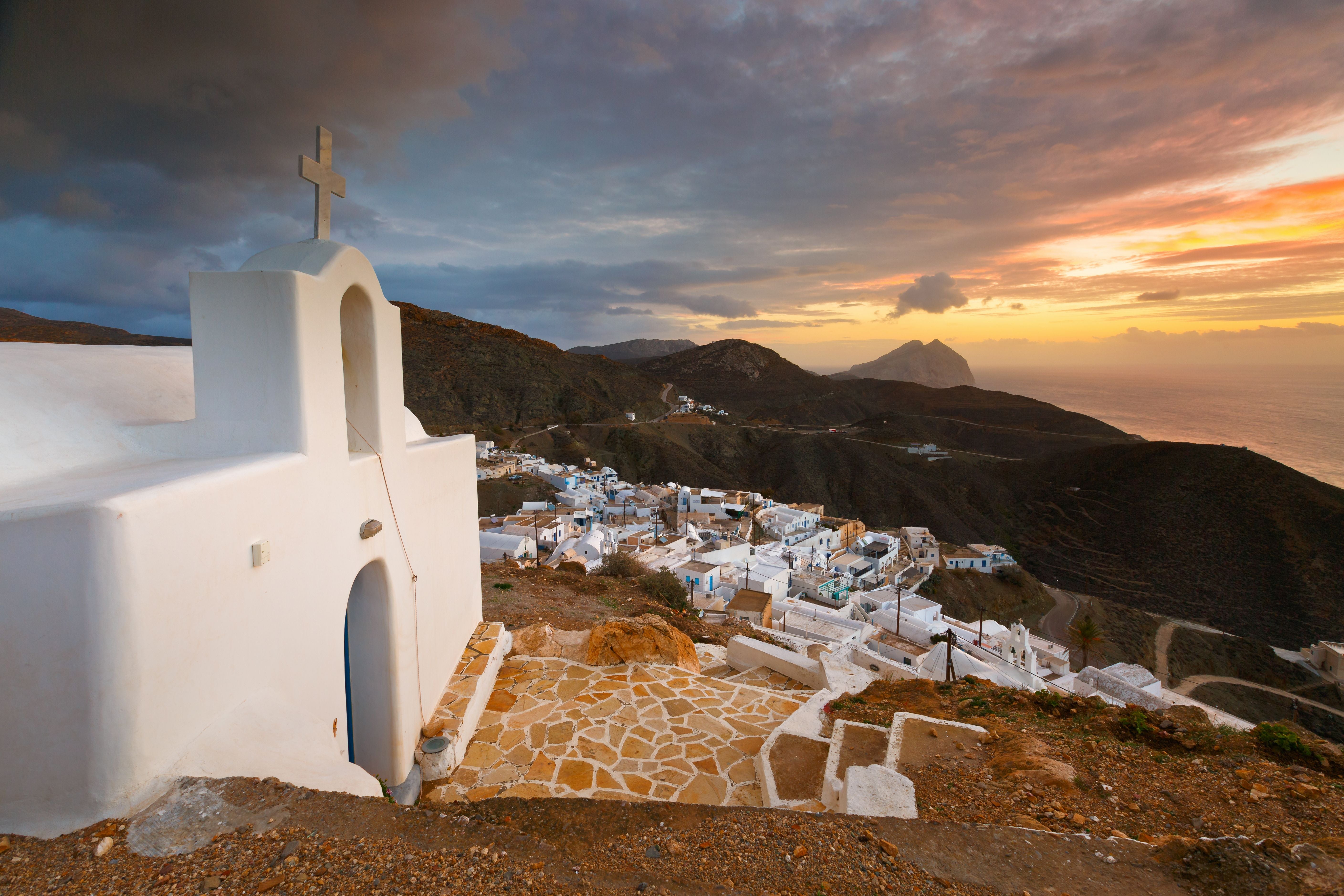 Weiße Kirche mit Kreuz im Vordergrund, griechisches Dorf und Meer im Sonnenuntergang.