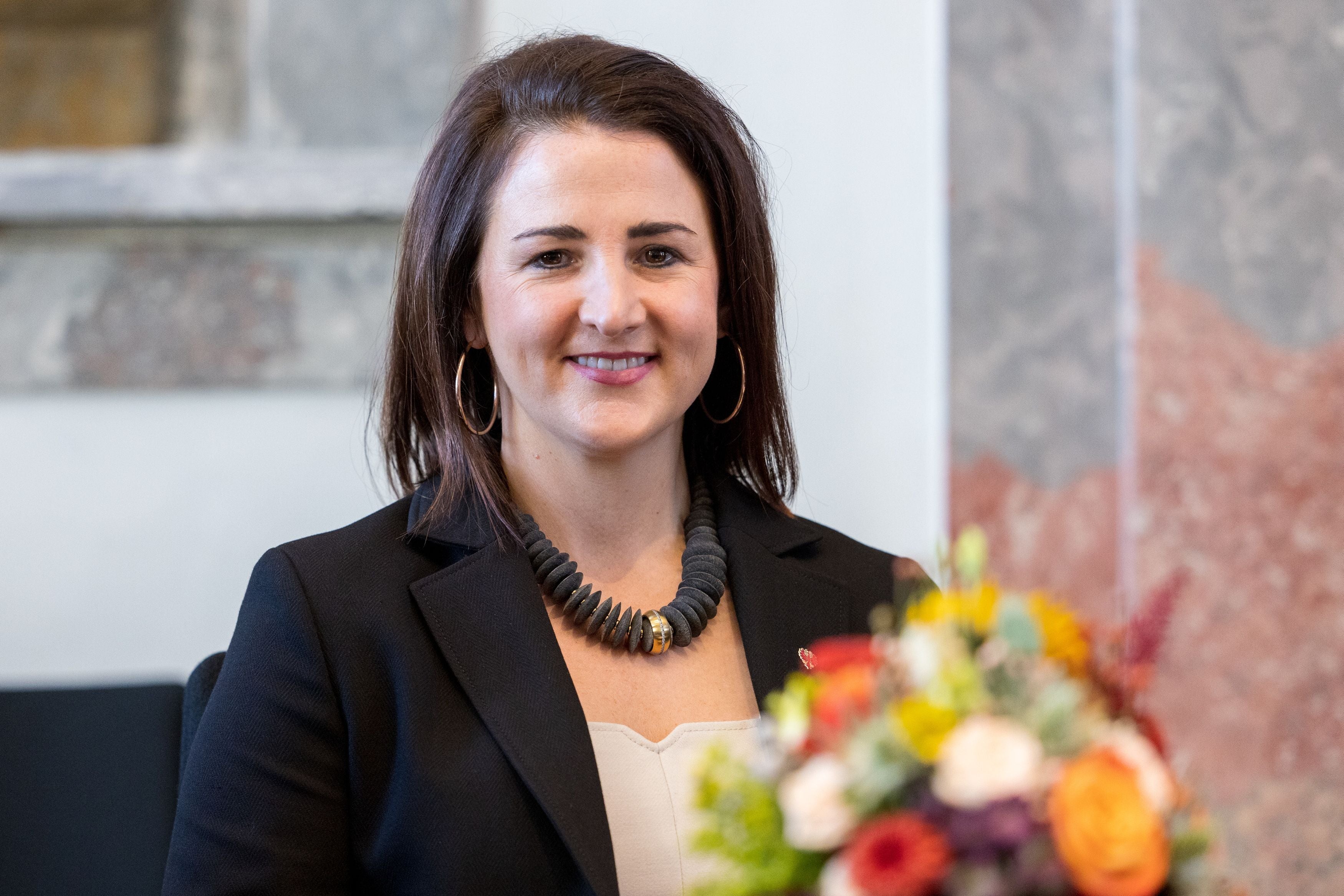 Eine Frau im dunklen Blazer sitzt lächelnd neben einem Blumenstrauß.