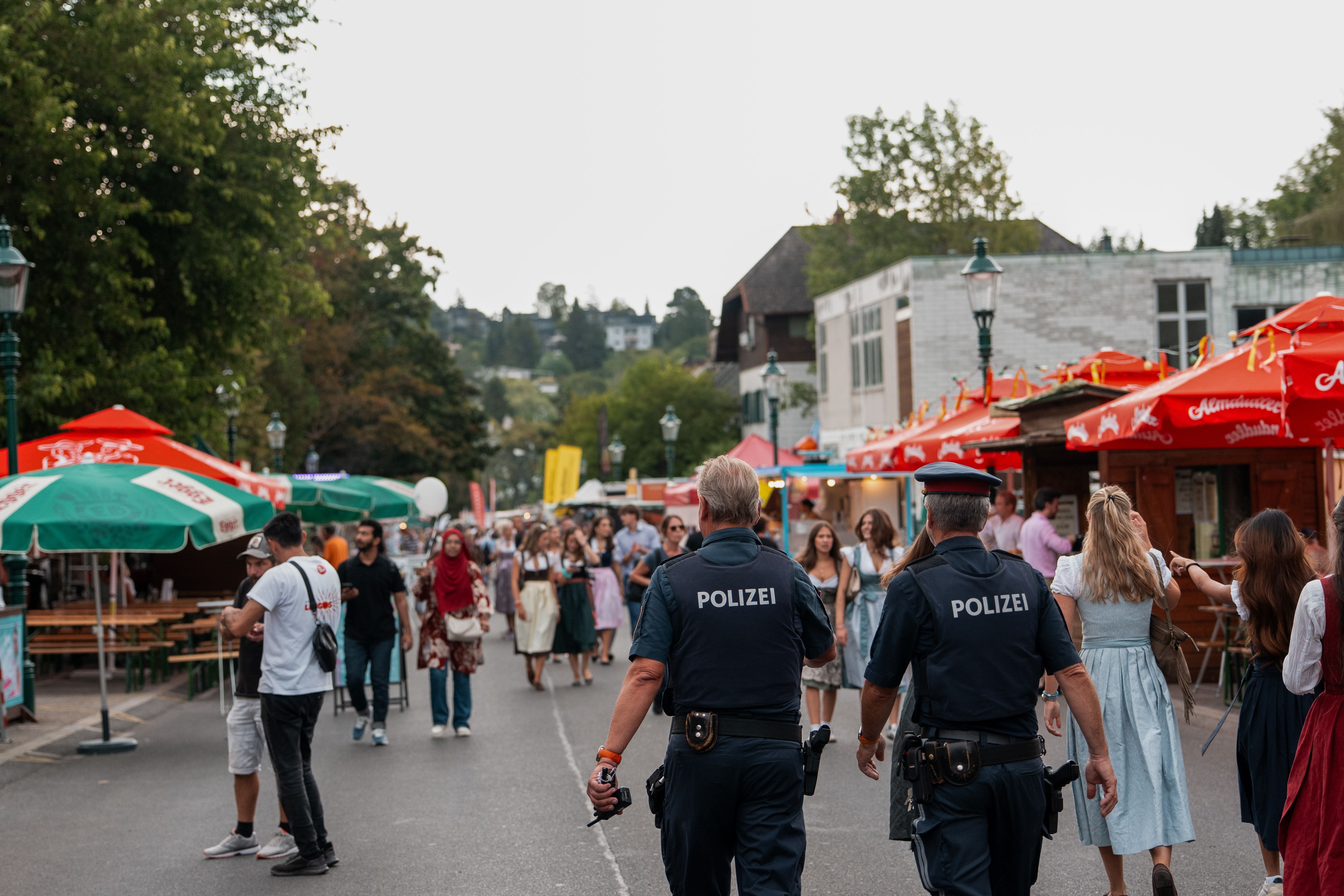 Zwei Polizisten gehen über ein belebtes Straßenfest mit bunten Sonnenschirmen in Wien.