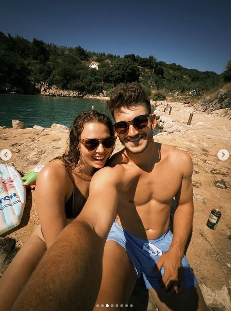 Ein junges Paar macht ein Selfie am felsigen Strand, beide tragen Sonnenbrillen und Badesachen.