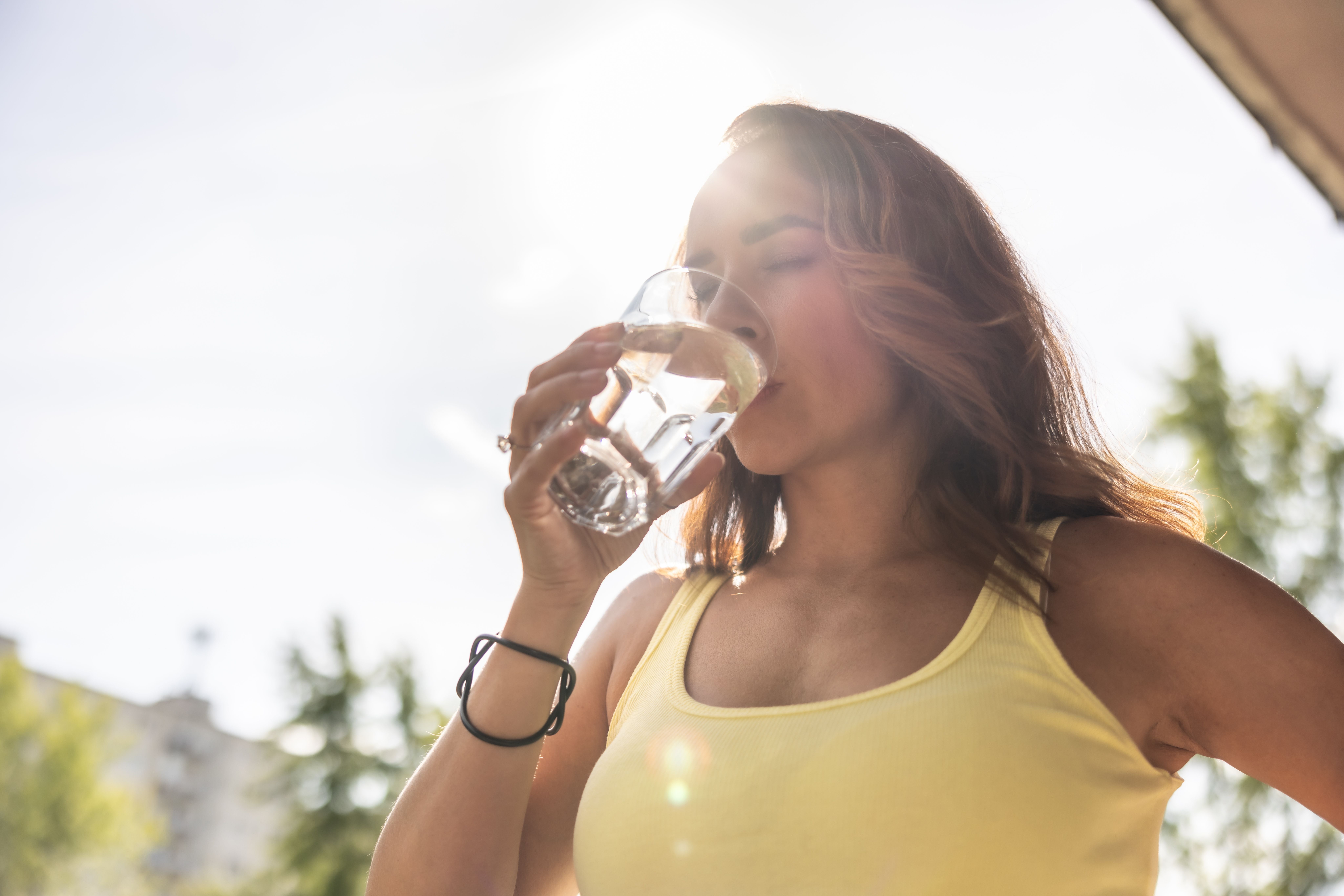 Frau in gelbem Top trinkt draußen an einem sonnigen Tag ein Glas Wasser.