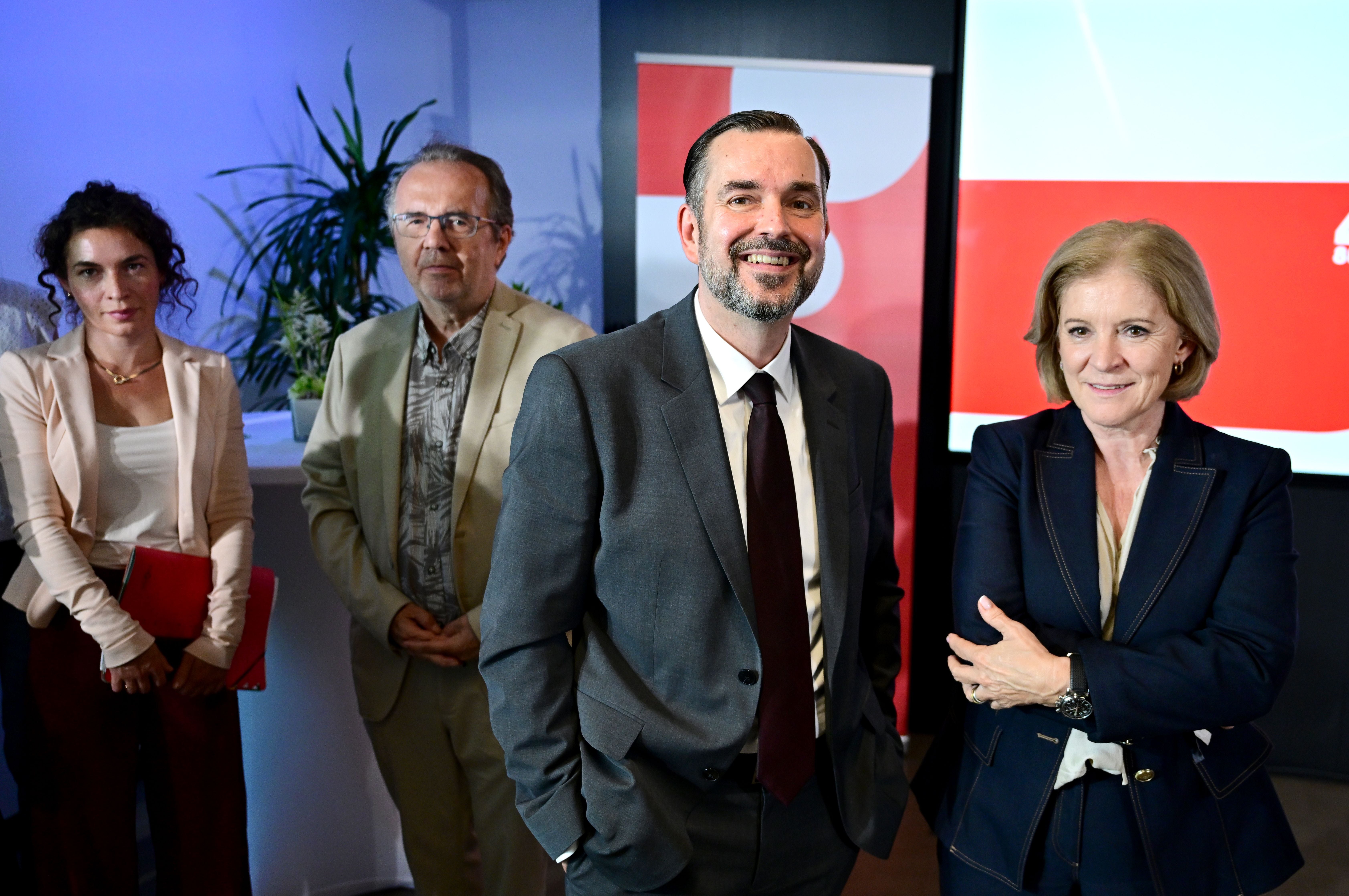 Vier Personen in Businesskleidung stehen bei einer Veranstaltung vor einem Präsentationsbildschirm.