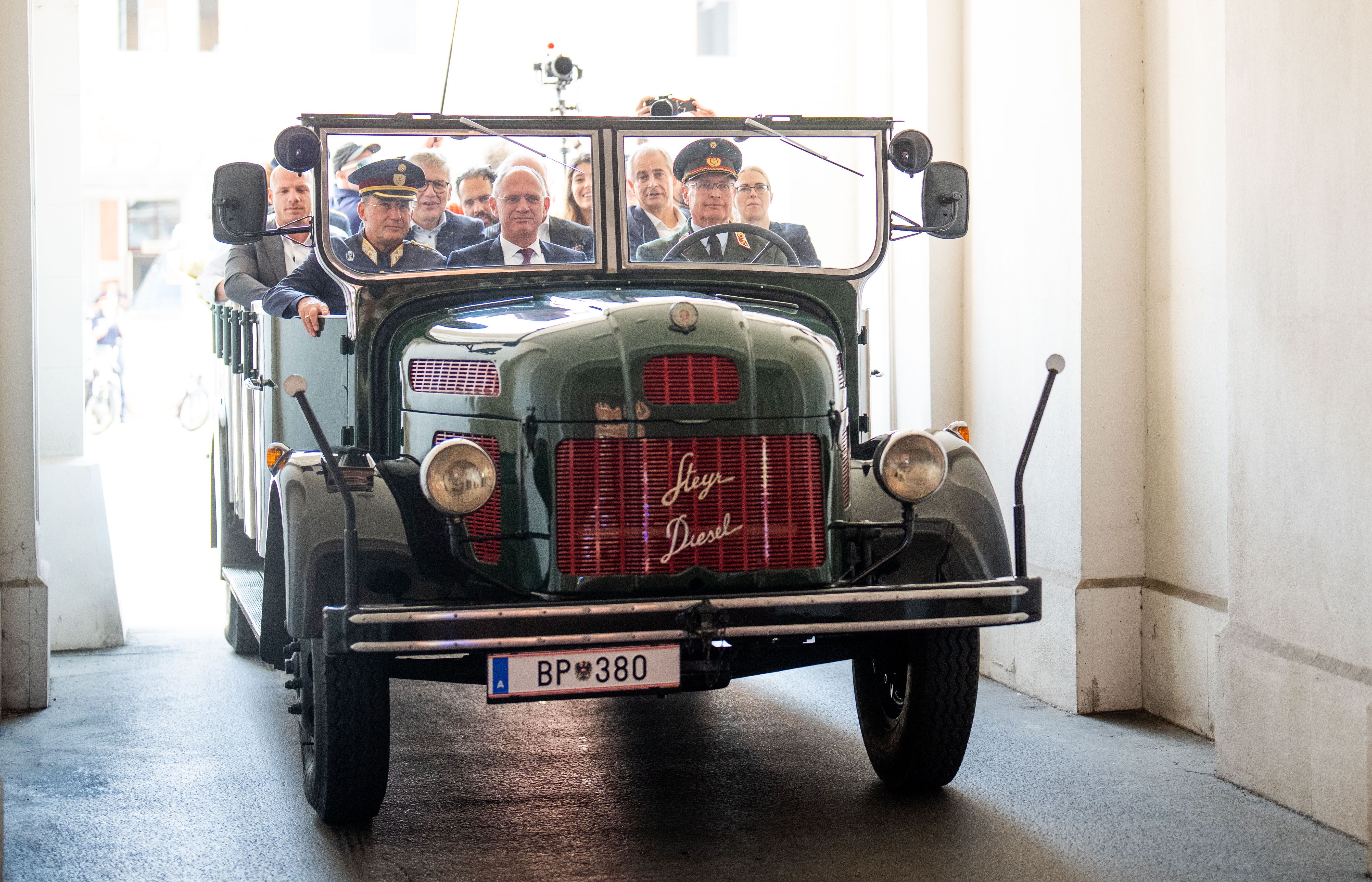 Historischer Polizeibus mit mehreren uniformierten und zivilen Personen fährt durch einen Torbogen.