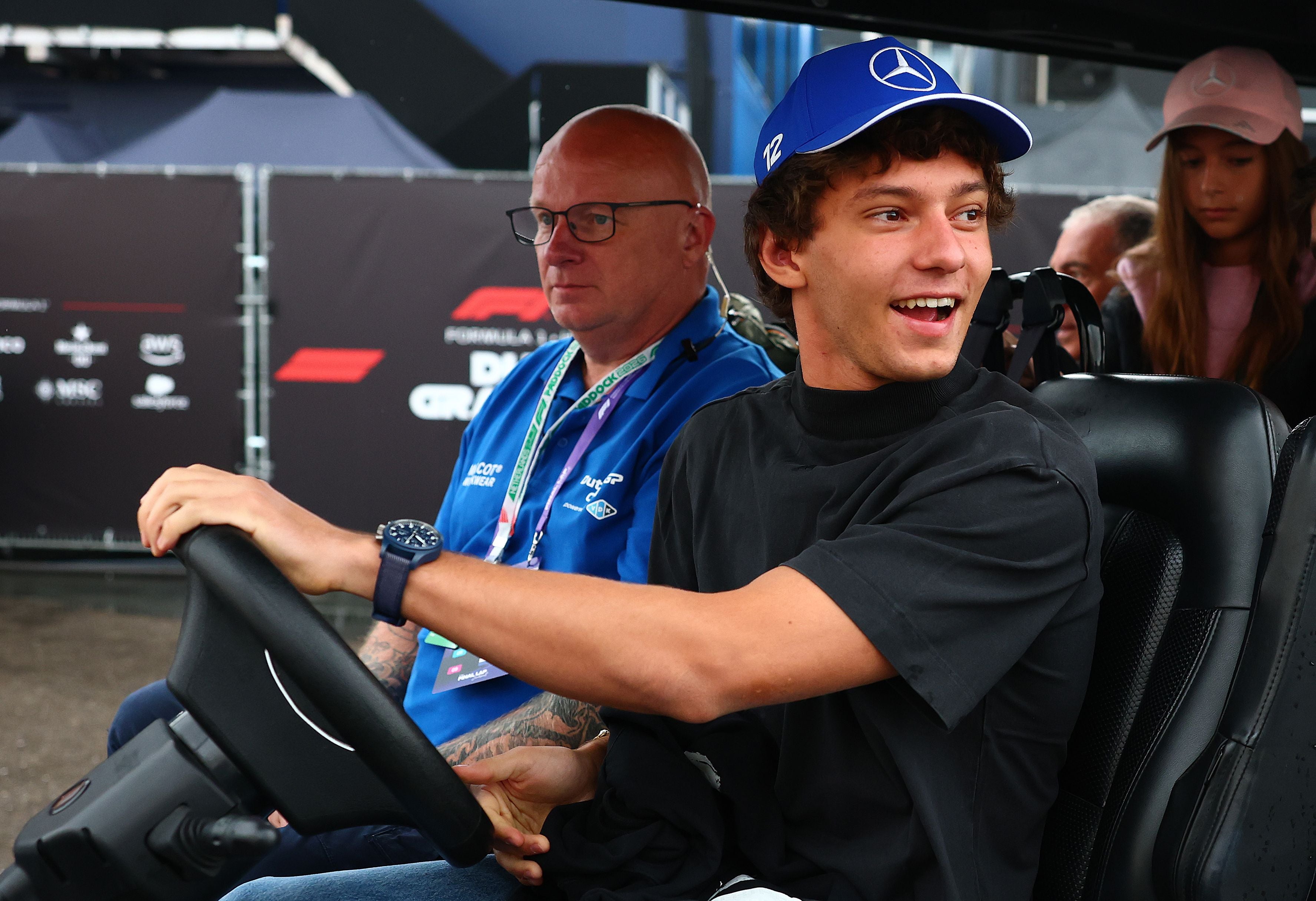 Ein junger Mann mit Mercedes-Mütze fährt fröhlich einen Golfkart im Fahrerlager.