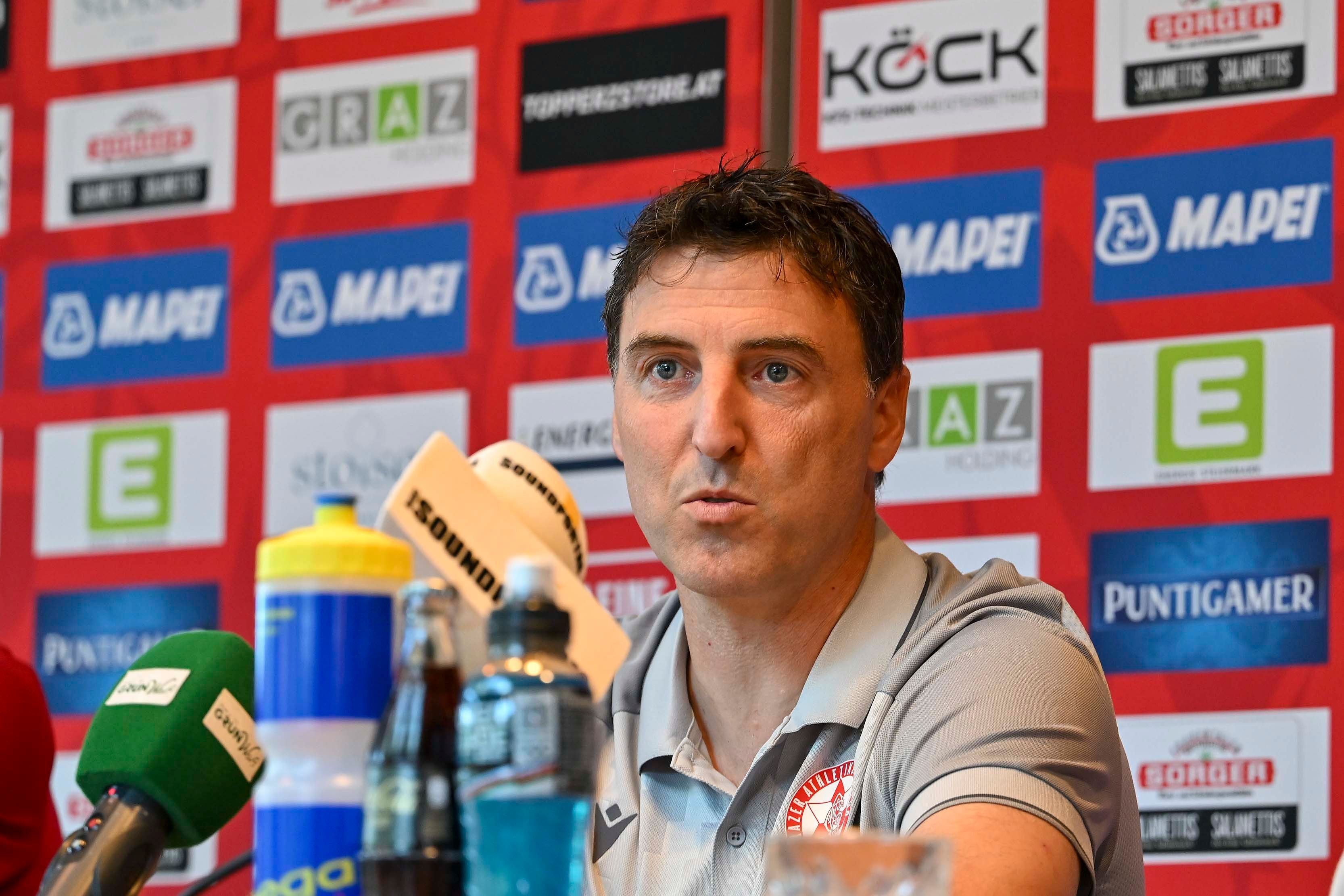 Ein Mann spricht auf einer Pressekonferenz vor Mikrofonen und Getränkeflaschen.