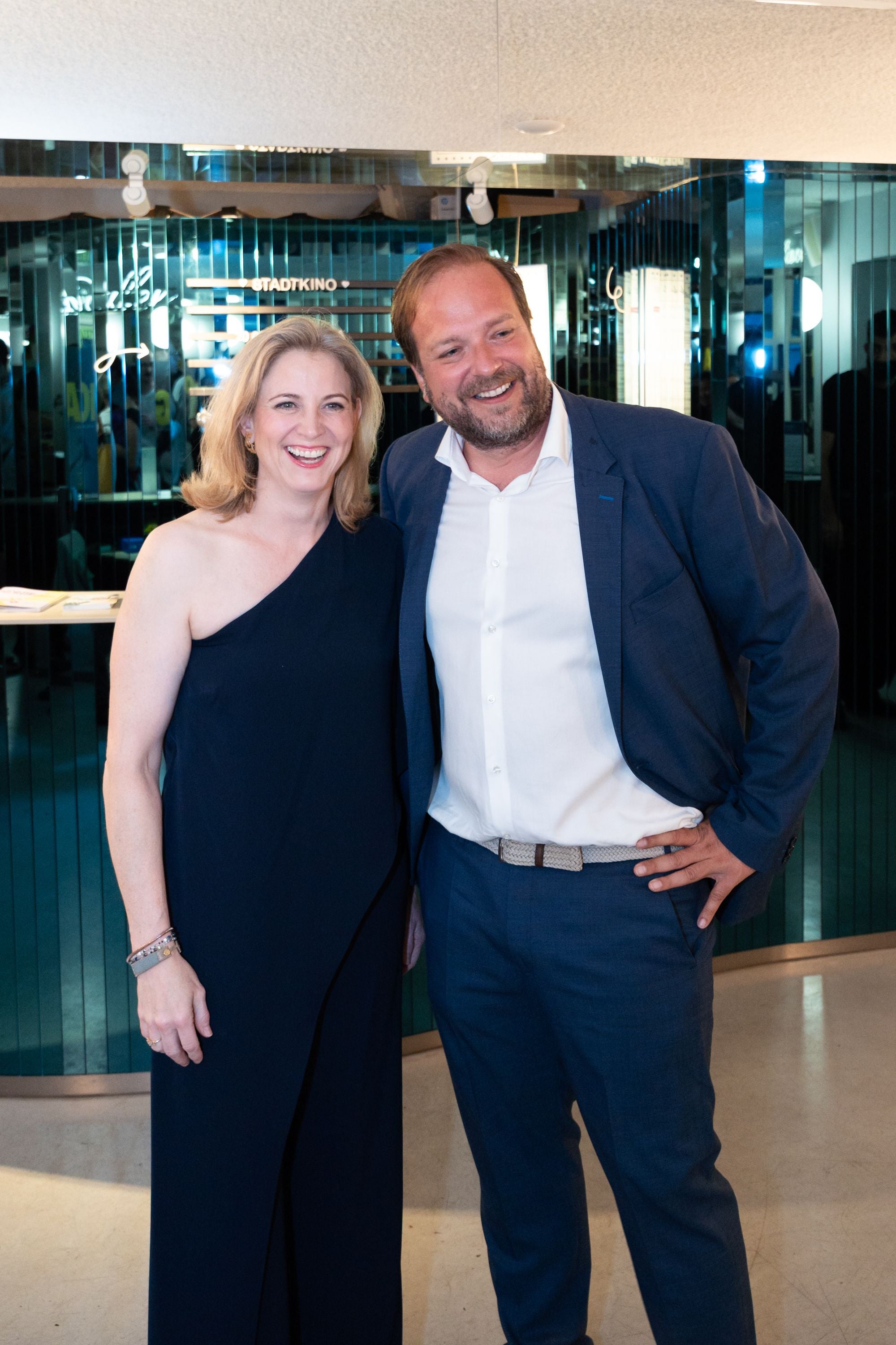 Eine Frau im schwarzen Kleid und ein Mann im blauen Anzug posieren lächelnd für ein Foto.