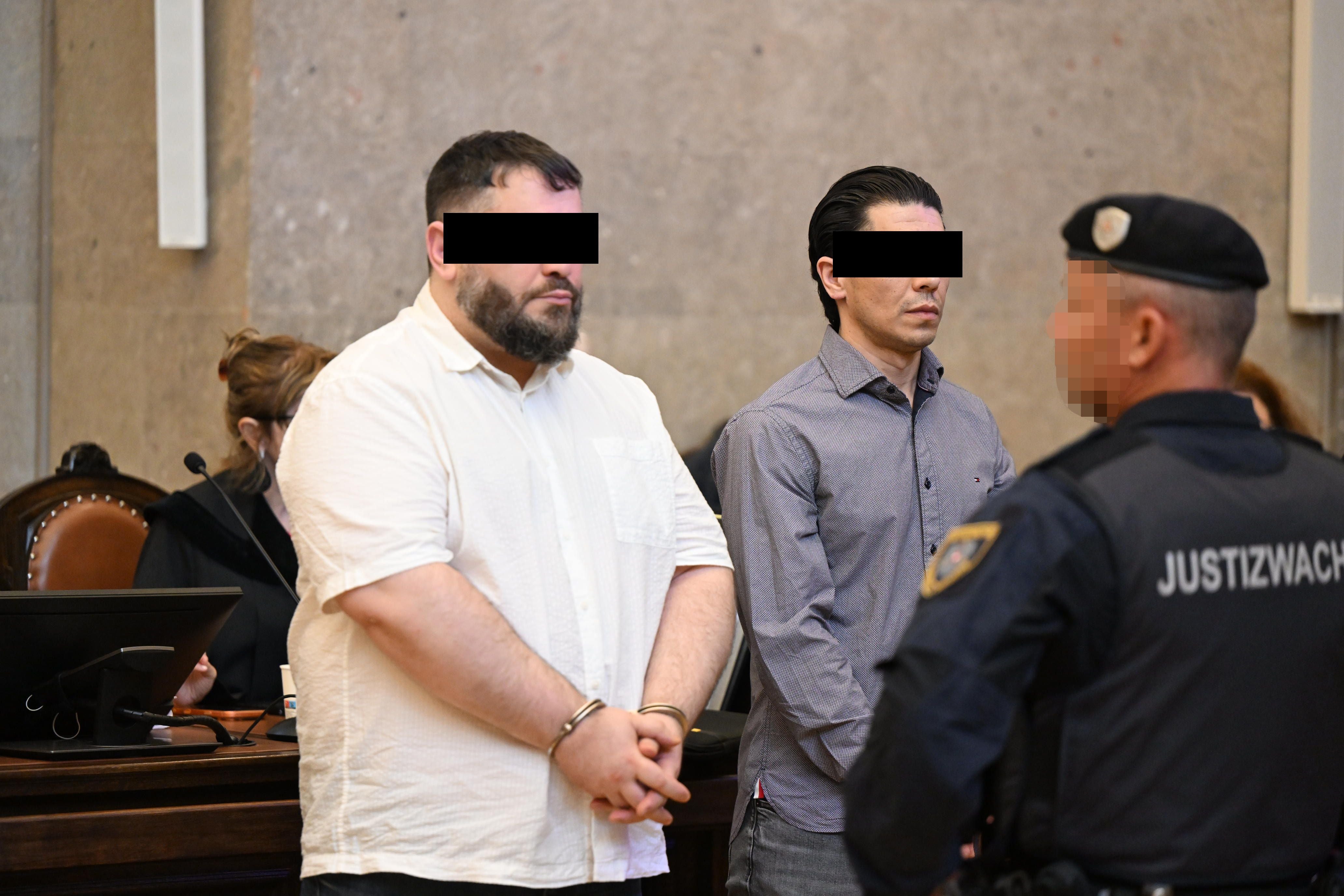 Zwei Angeklagte mit schwarzen Balken vor den Augen stehen im Gericht neben einem Justizwachebeamten.