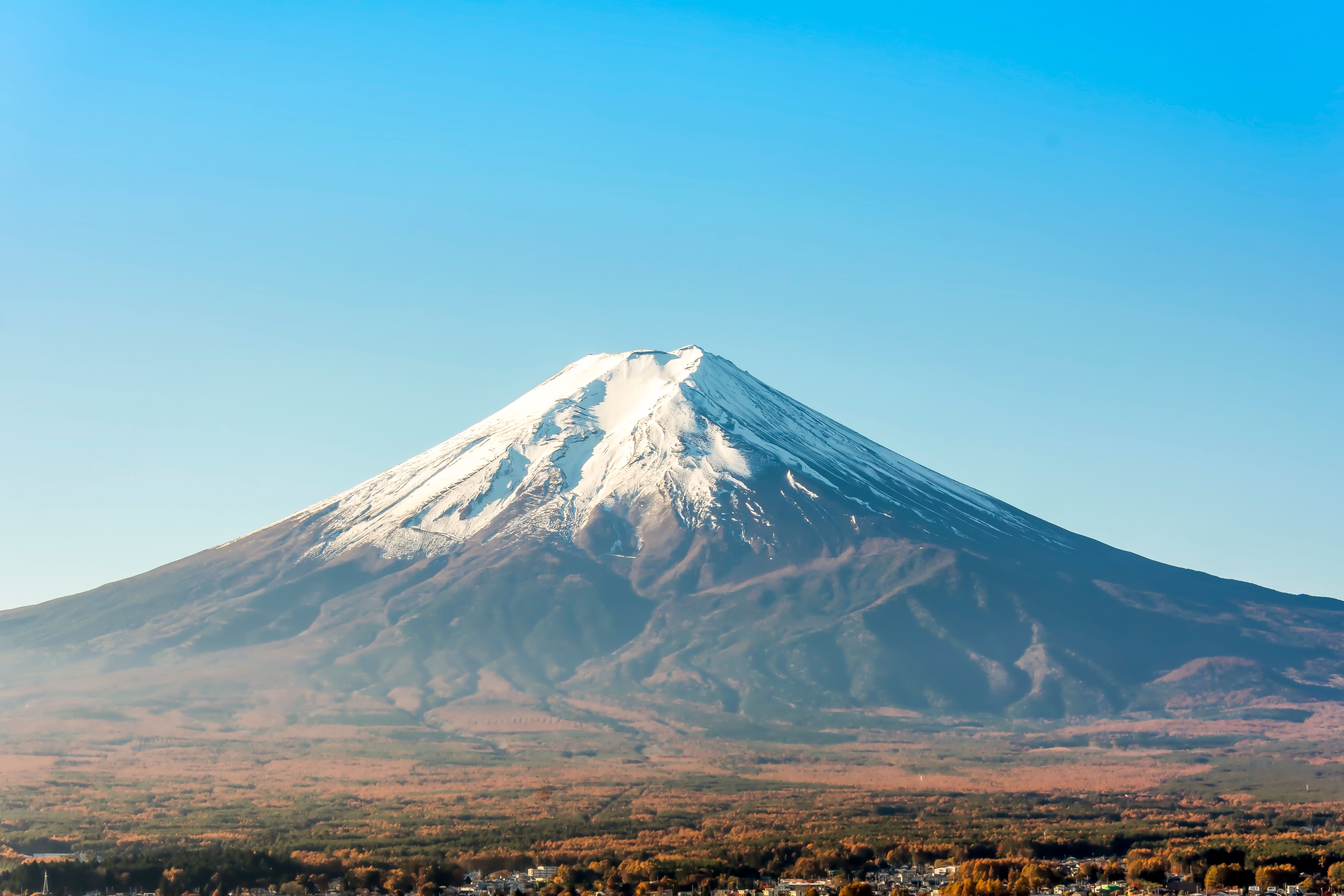 Schneebedeckter Vulkan Fuji vor klarem, blauem Himmel mit herbstlicher Landschaft im Vordergrund.