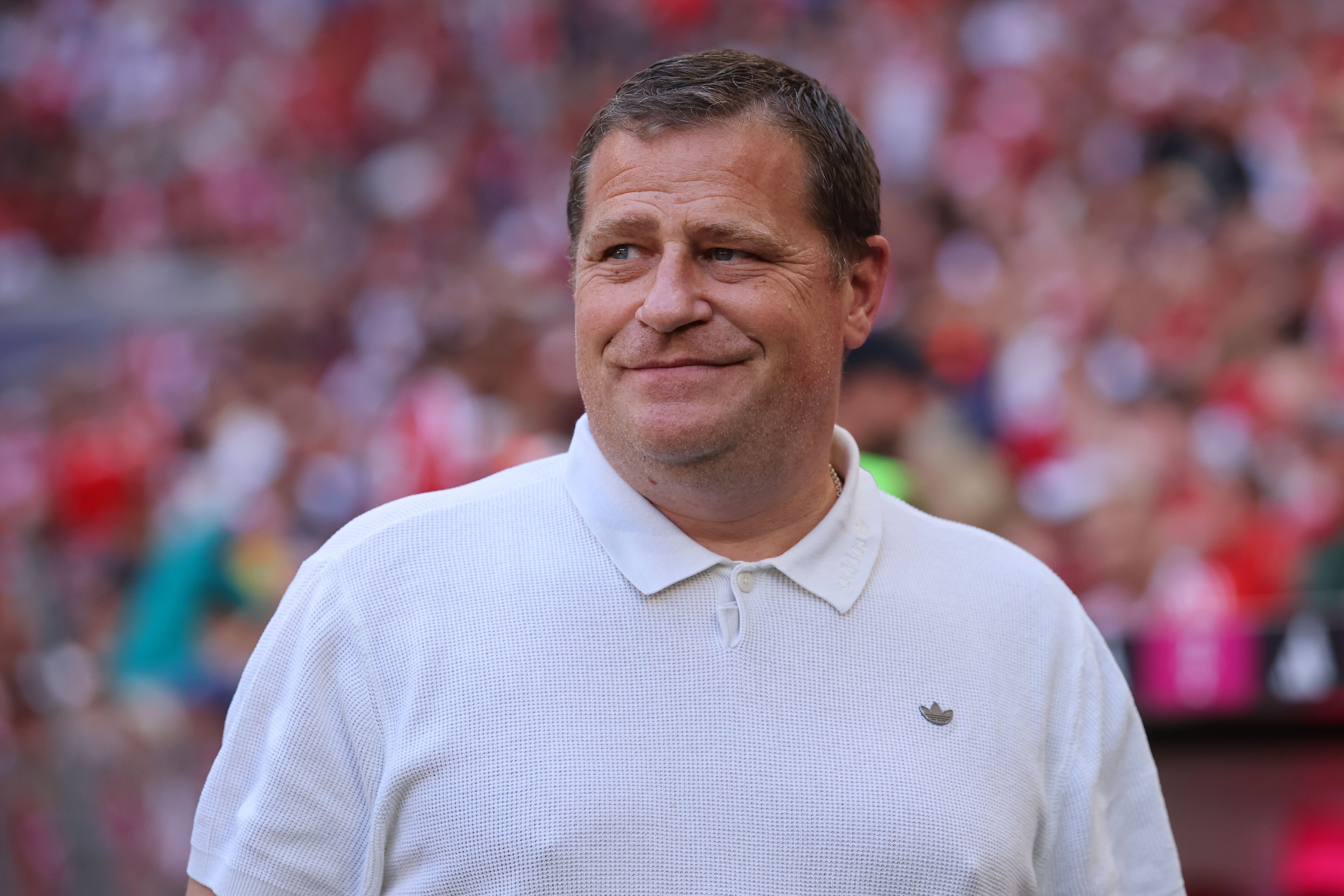Ein Mann in weißem Poloshirt steht lächelnd in einem Stadion mit unscharfem Publikum.