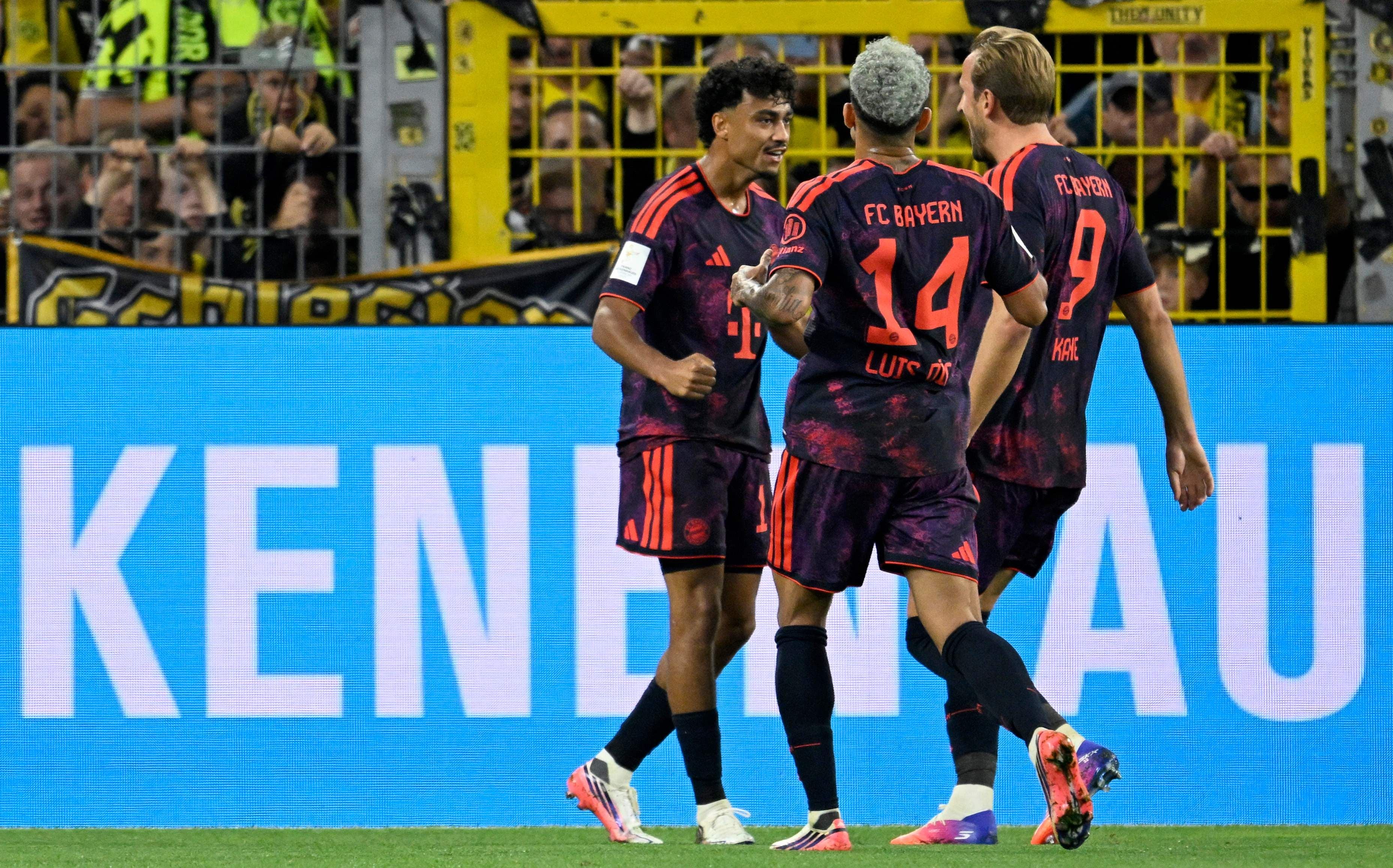 Drei Bayern-Spieler feiern ein Tor im Stadion vor jubelnden Fans im Hintergrund.
