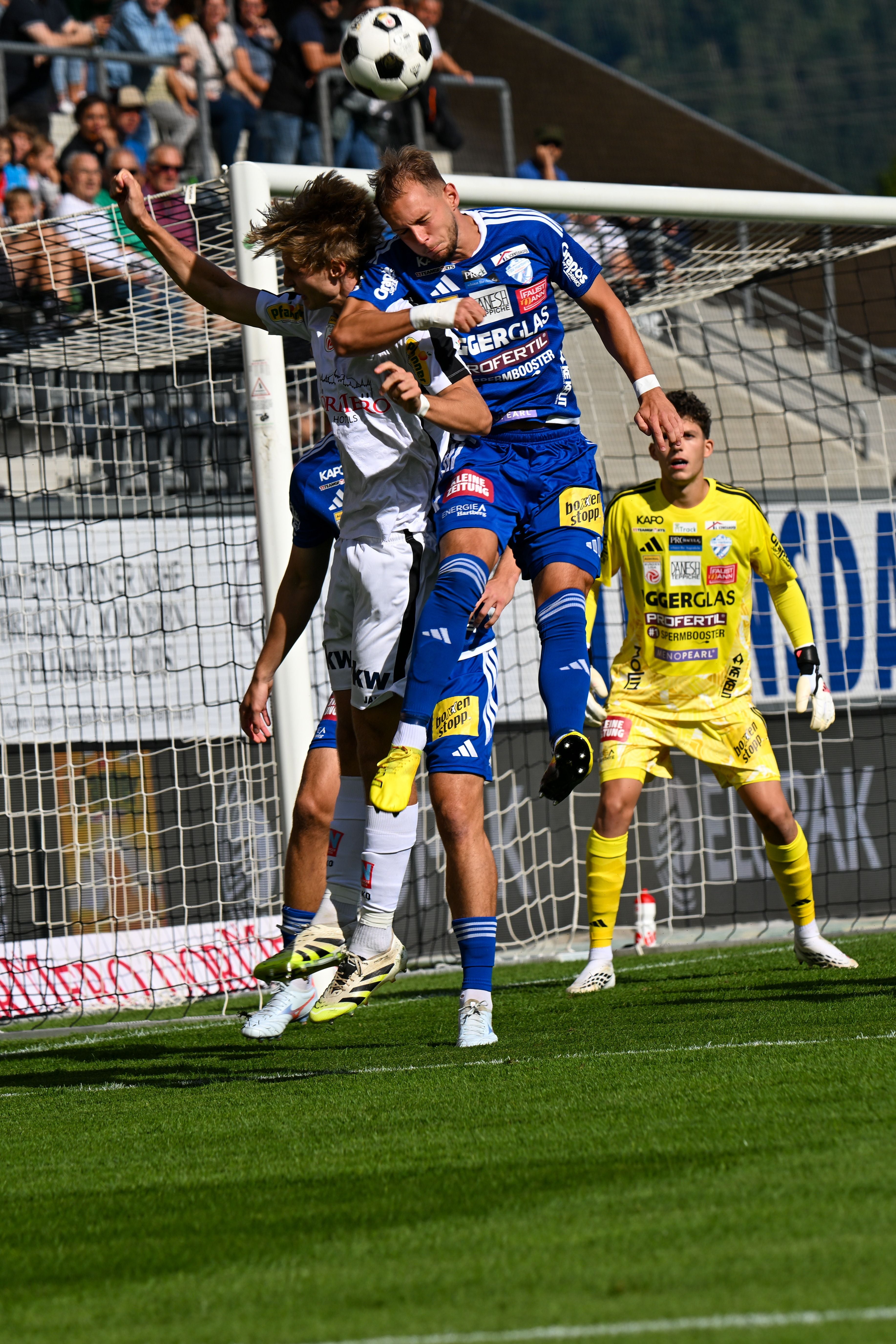 Zwei Fußballspieler springen zum Kopfball vor dem Tor, ein Torwart steht im Hintergrund.