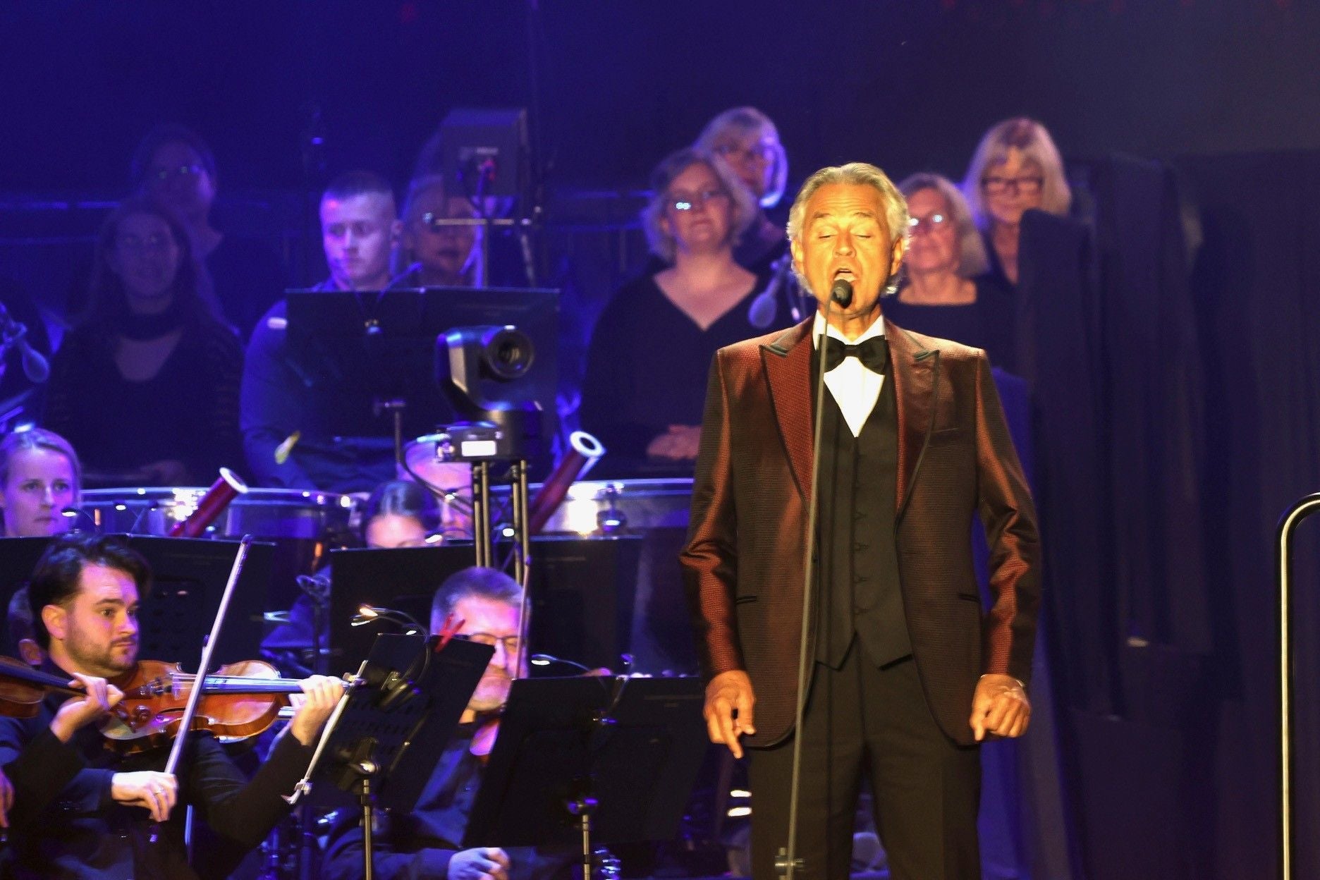 Andrea Bocelli singt im Anzug auf einer Bühne, begleitet von Orchester und Chor.