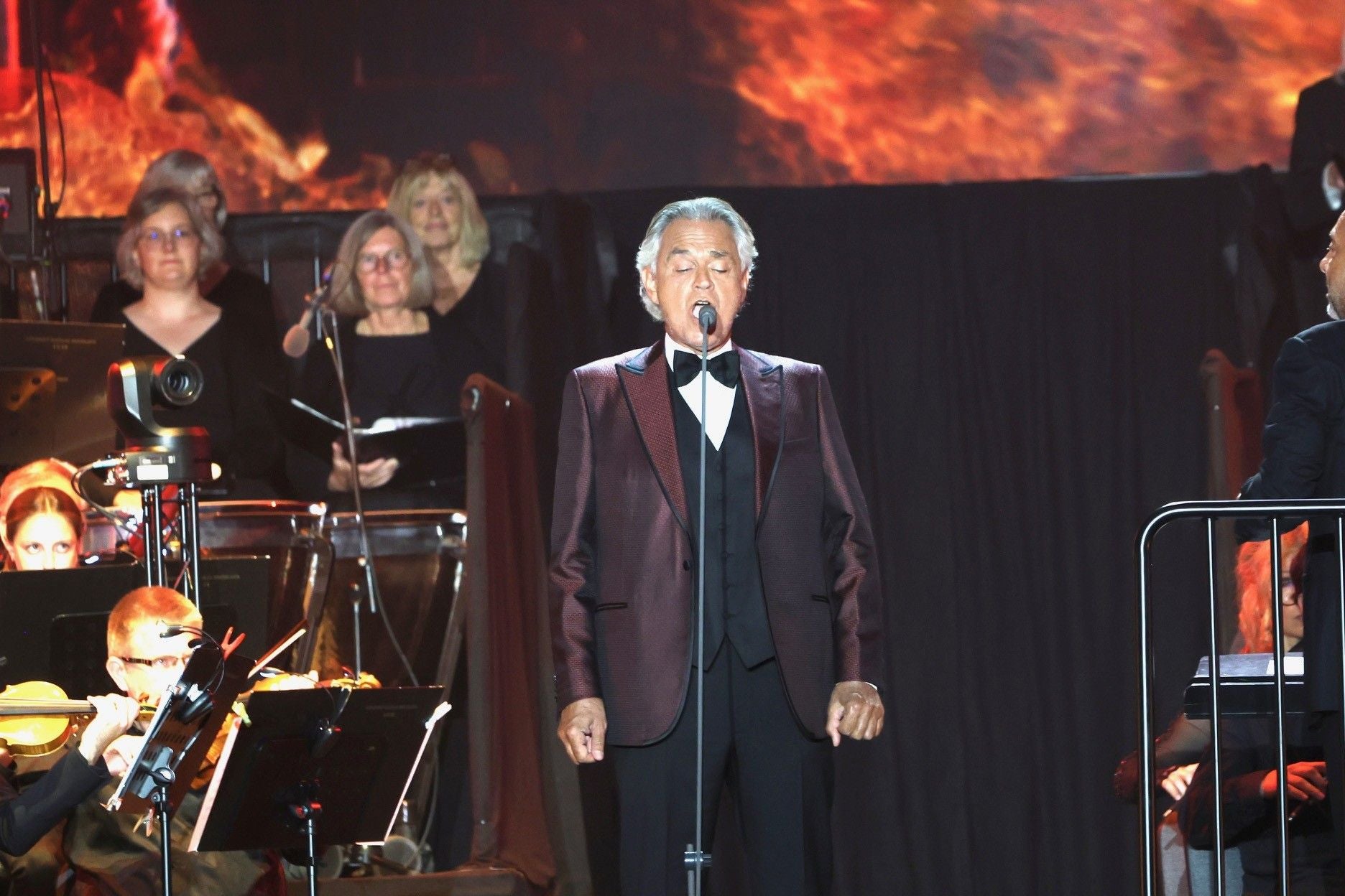 Andrea Bocelli singt mit geschlossenem Mund auf einer Bühne vor einem Orchester und Chor.