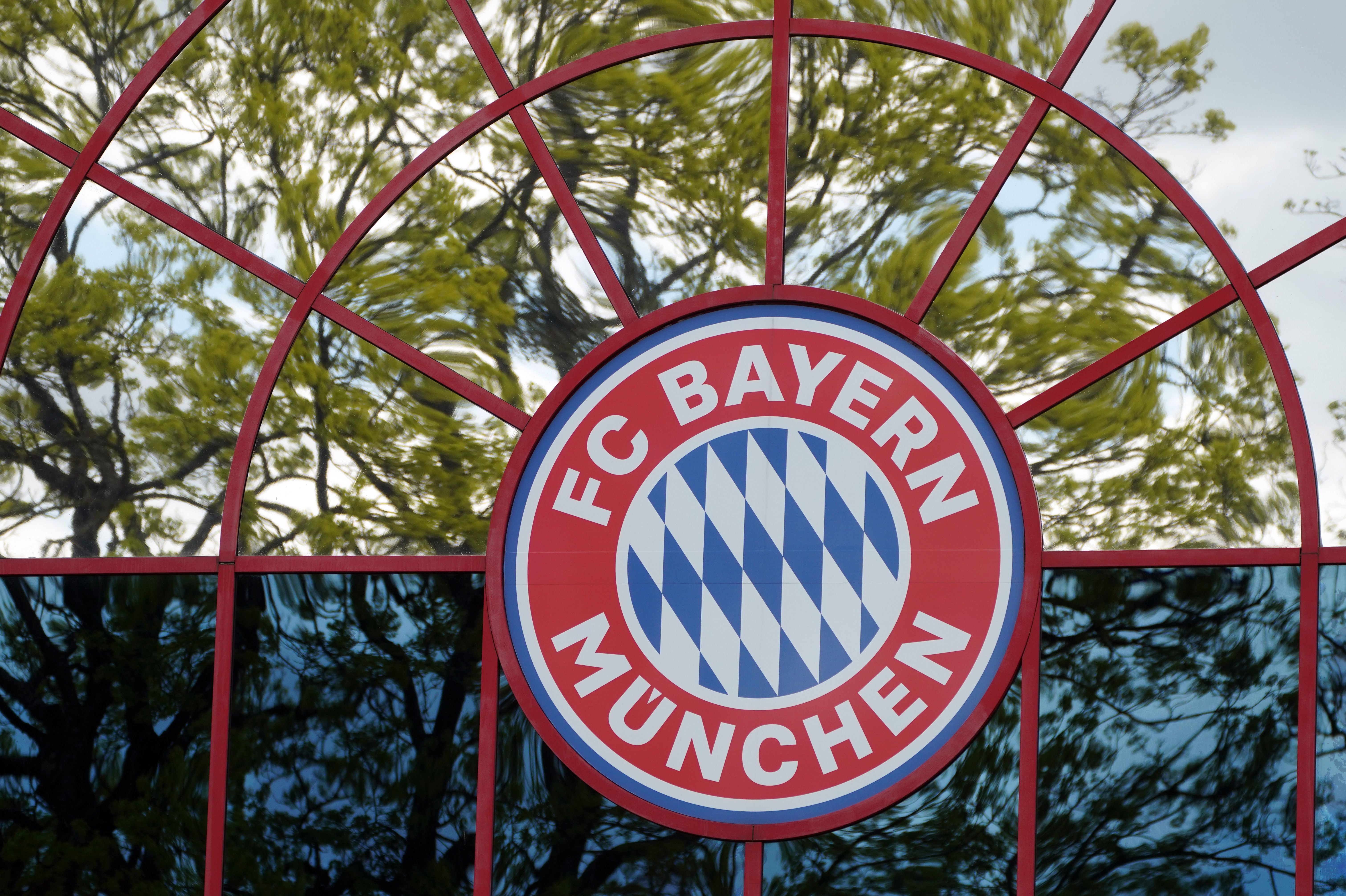 Das FC Bayern München-Logo an einem Fenster mit spiegelnden Bäumen im Hintergrund.