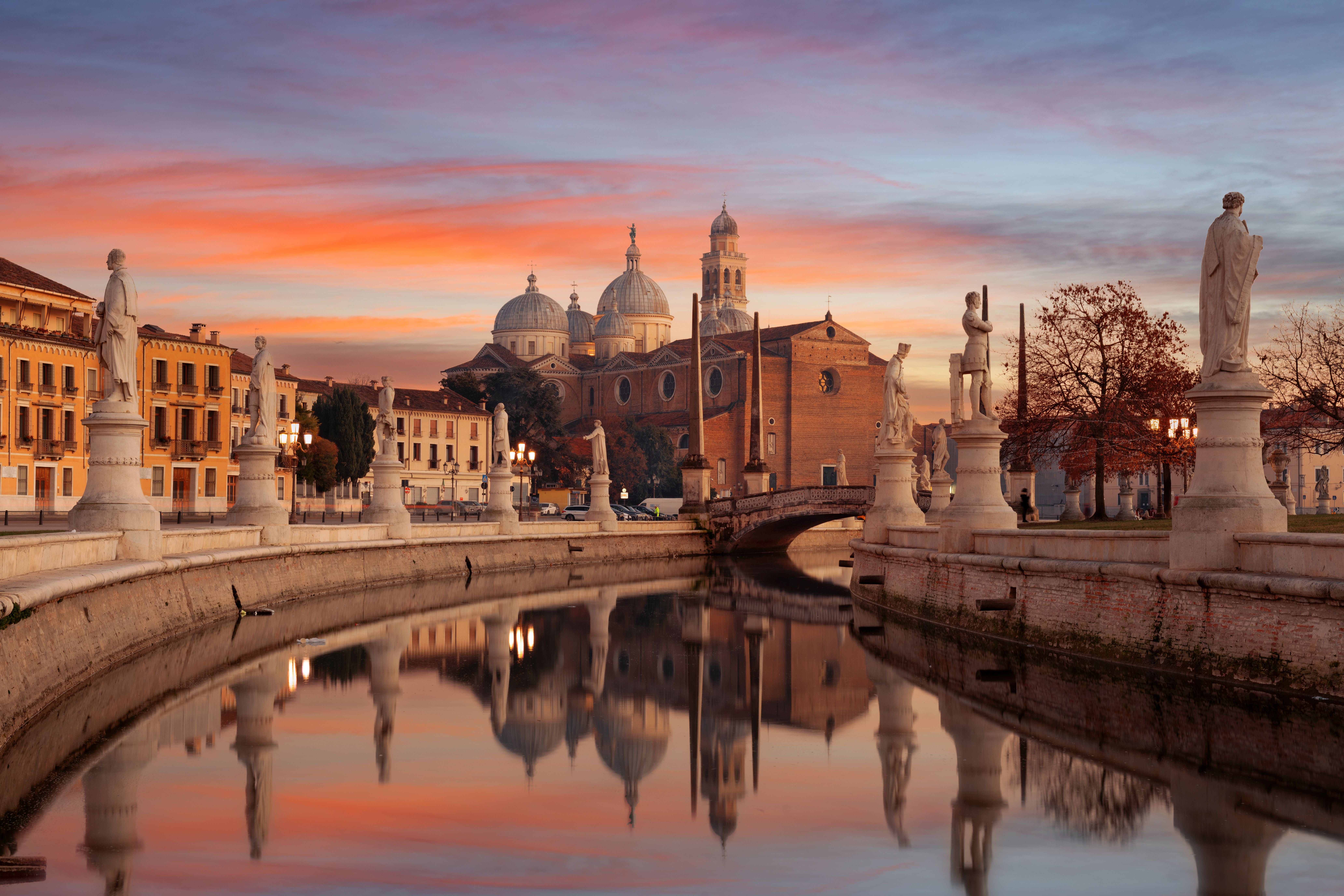 Prato della Valle in Padua mit Statuen und Kirche bei Sonnenuntergang, spiegelnd im Wasser.