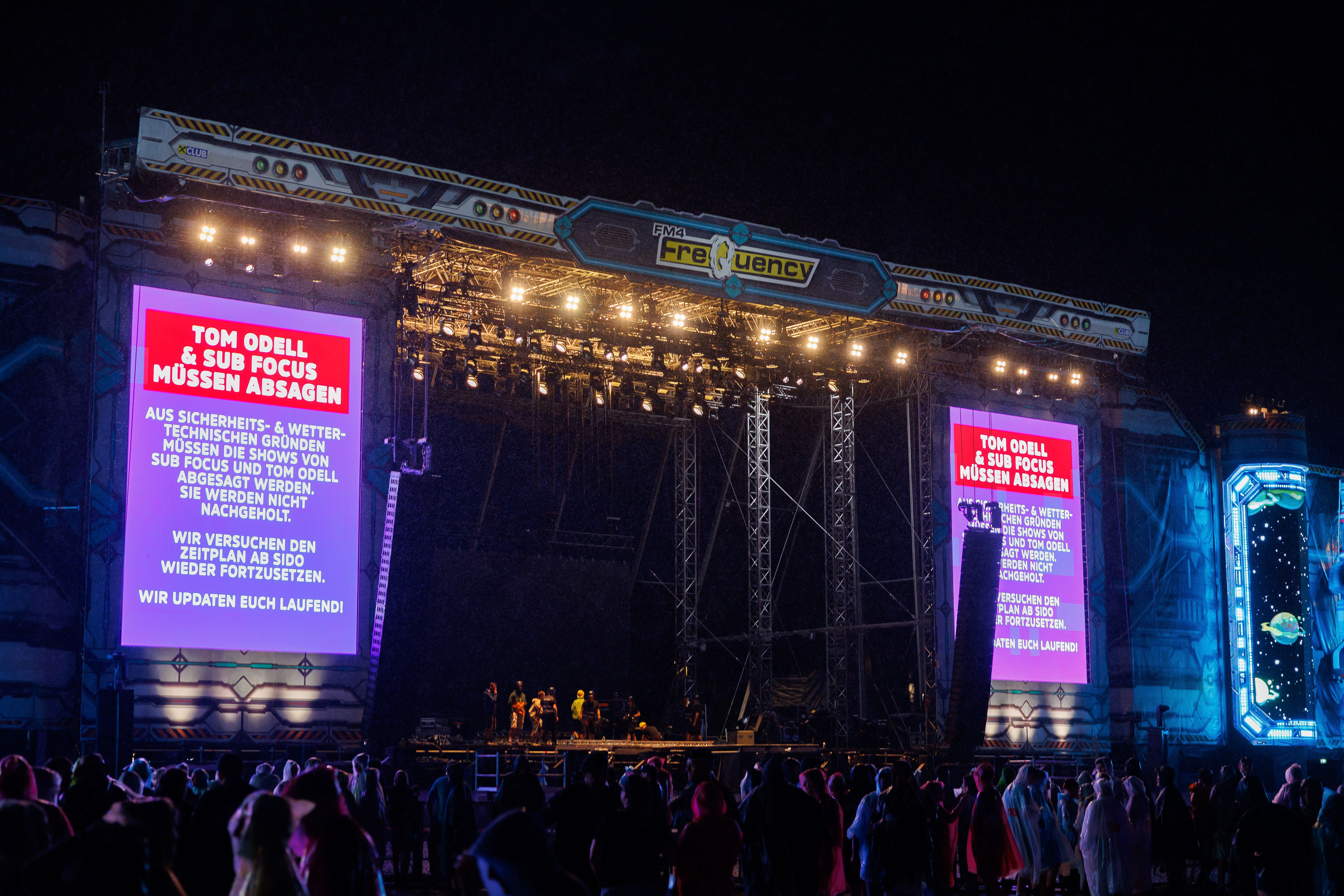 Menschen stehen vor der Frequency-Bühne, Monitore zeigen Absage von Tom Odell und Sub Focus wegen Sturm.