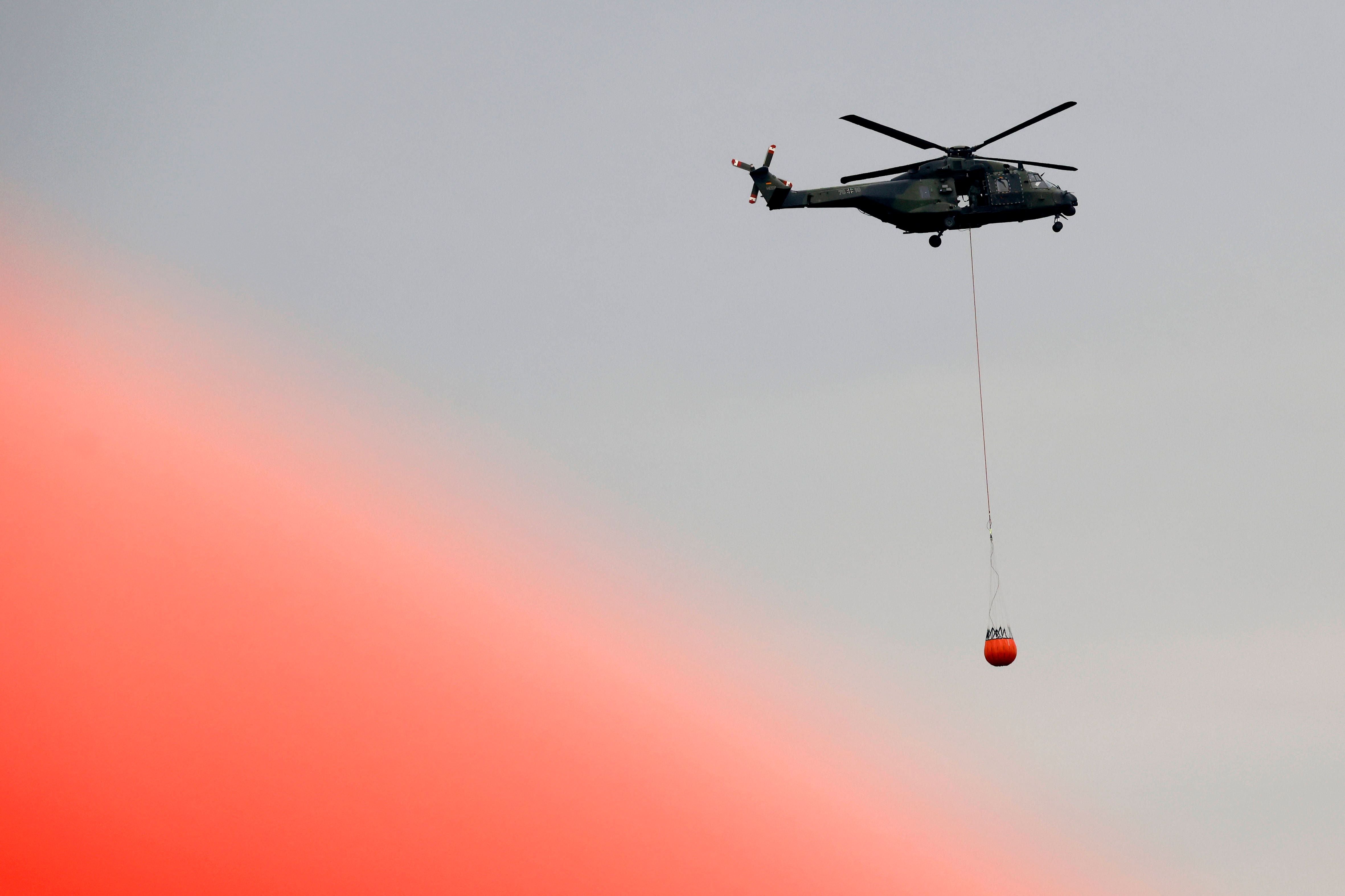 Ein Hubschrauber mit Löschwasserbehälter fliegt über einen rauchigen, bewölkten Himmel.