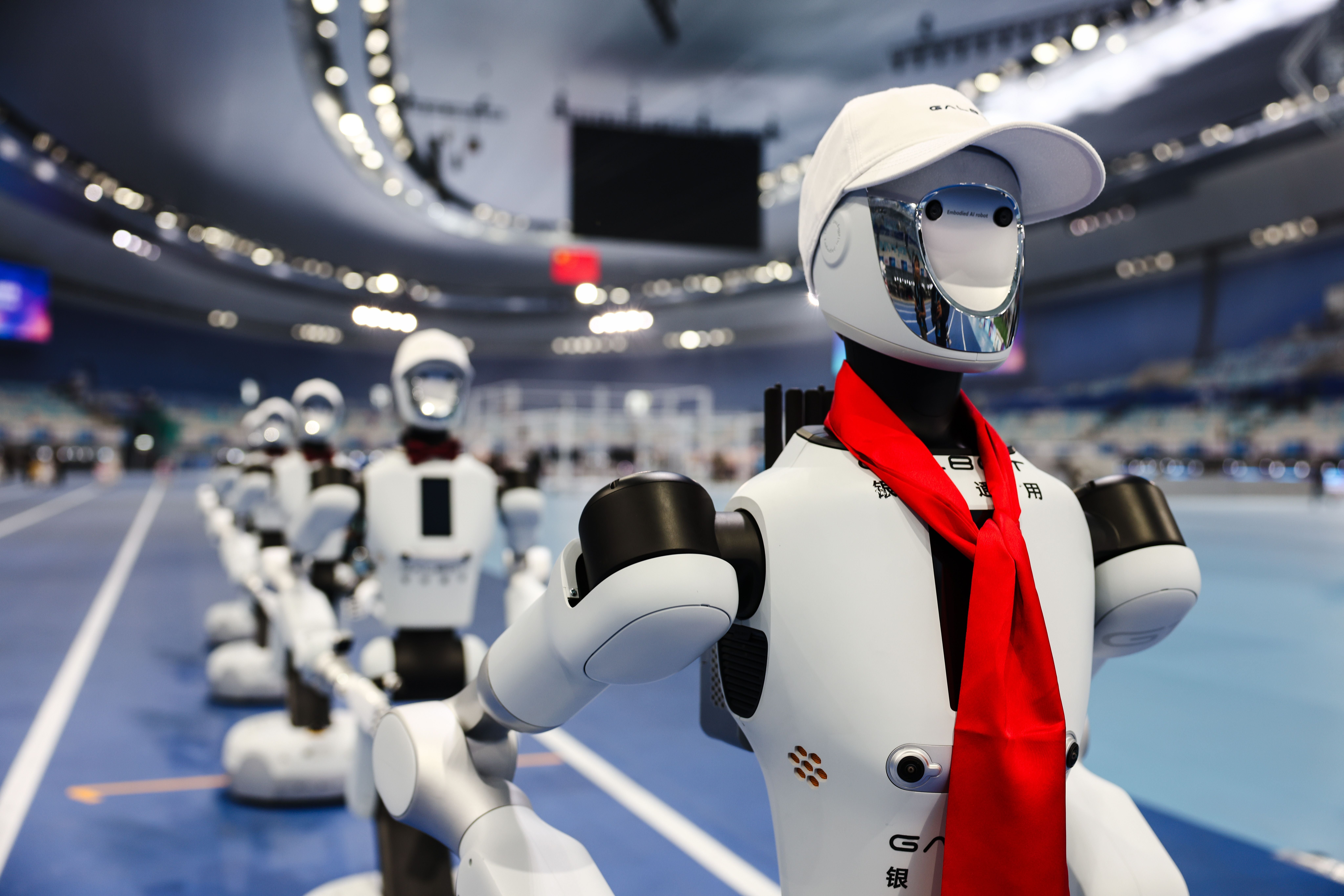 Mehrere humanoide Roboter mit roten Halstüchern stehen in einer Sportarena in Peking.