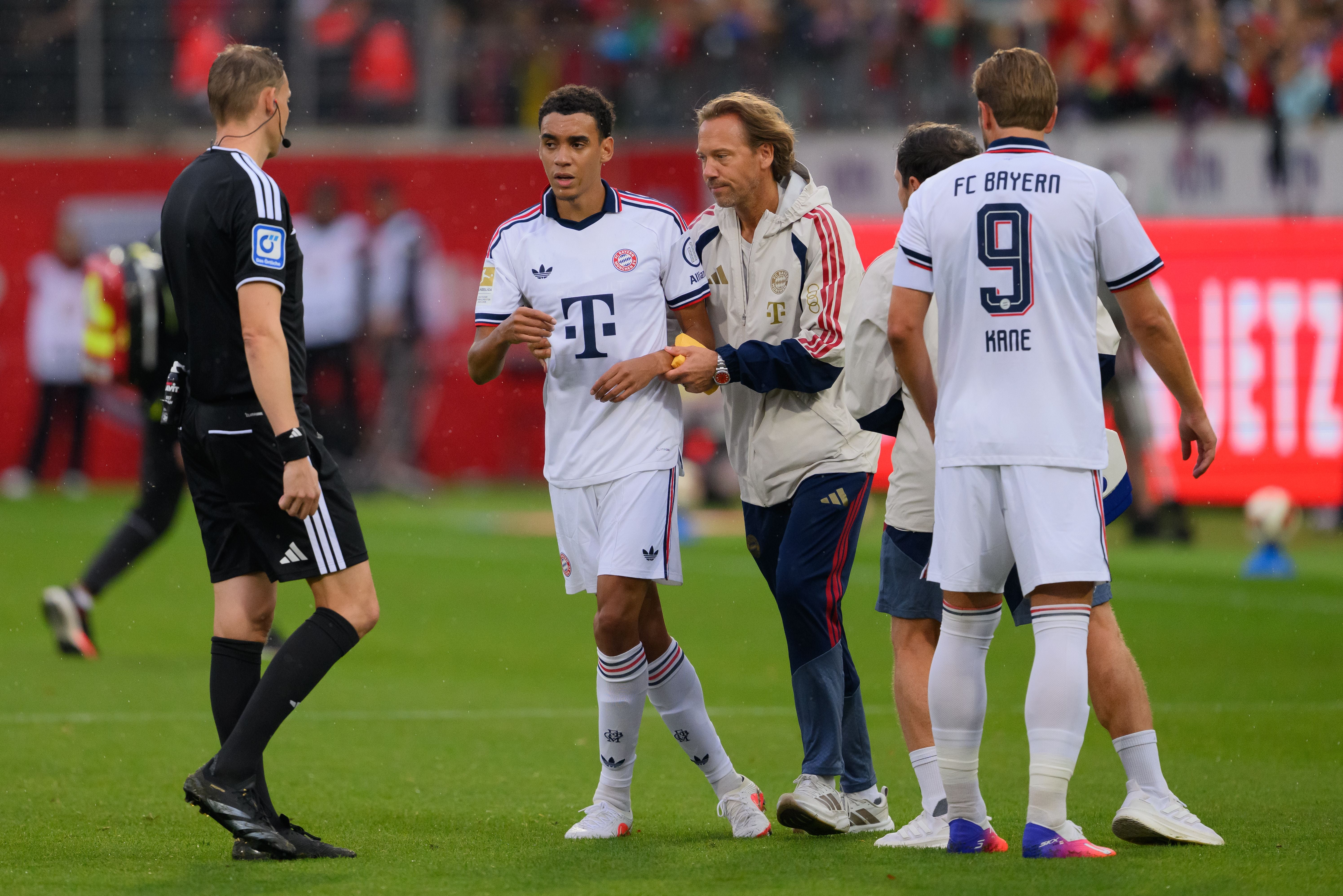 Jamal Musiala verlässt mit Unterstützung das Spielfeld, neben ihm Mitspieler und ein Schiedsrichter.