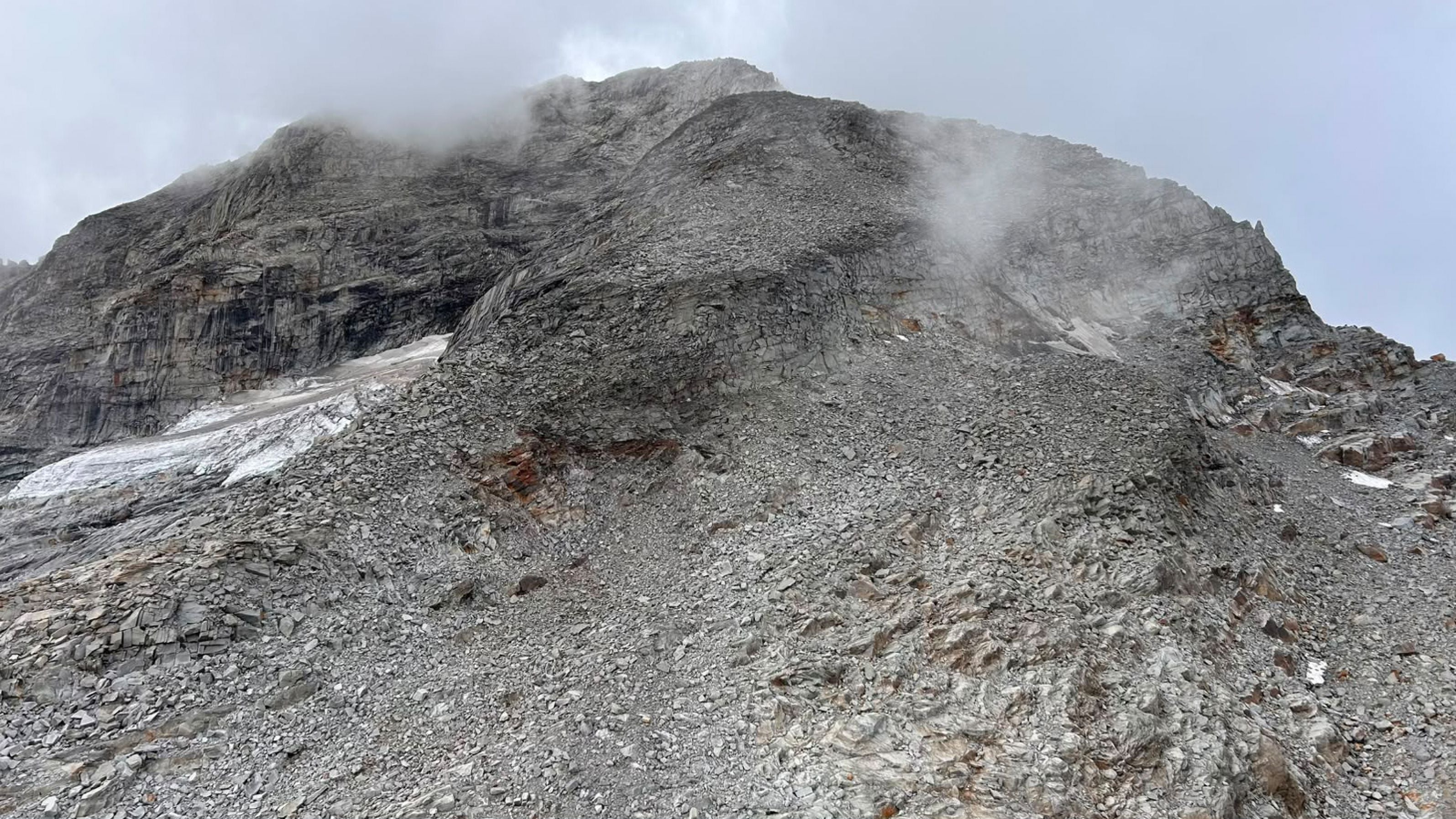 Steiniger Berghang mit Nebel und vereinzelten Schneeresten unter bewölktem Himmel.