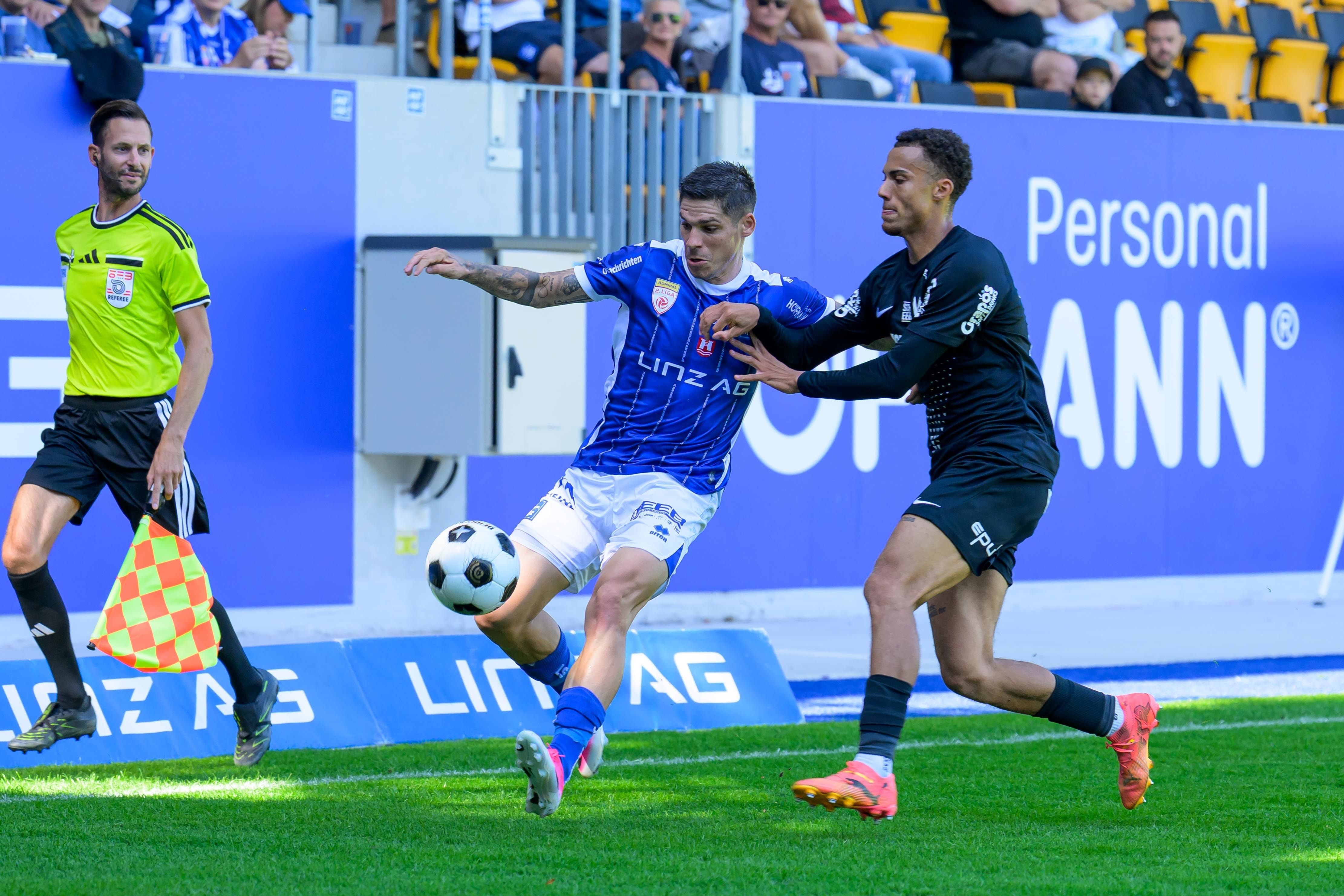 Zwei Fußballspieler kämpfen auf dem Rasen um den Ball, während der Schiedsrichterassistent mit der Linienrichterfahne daneben steht.