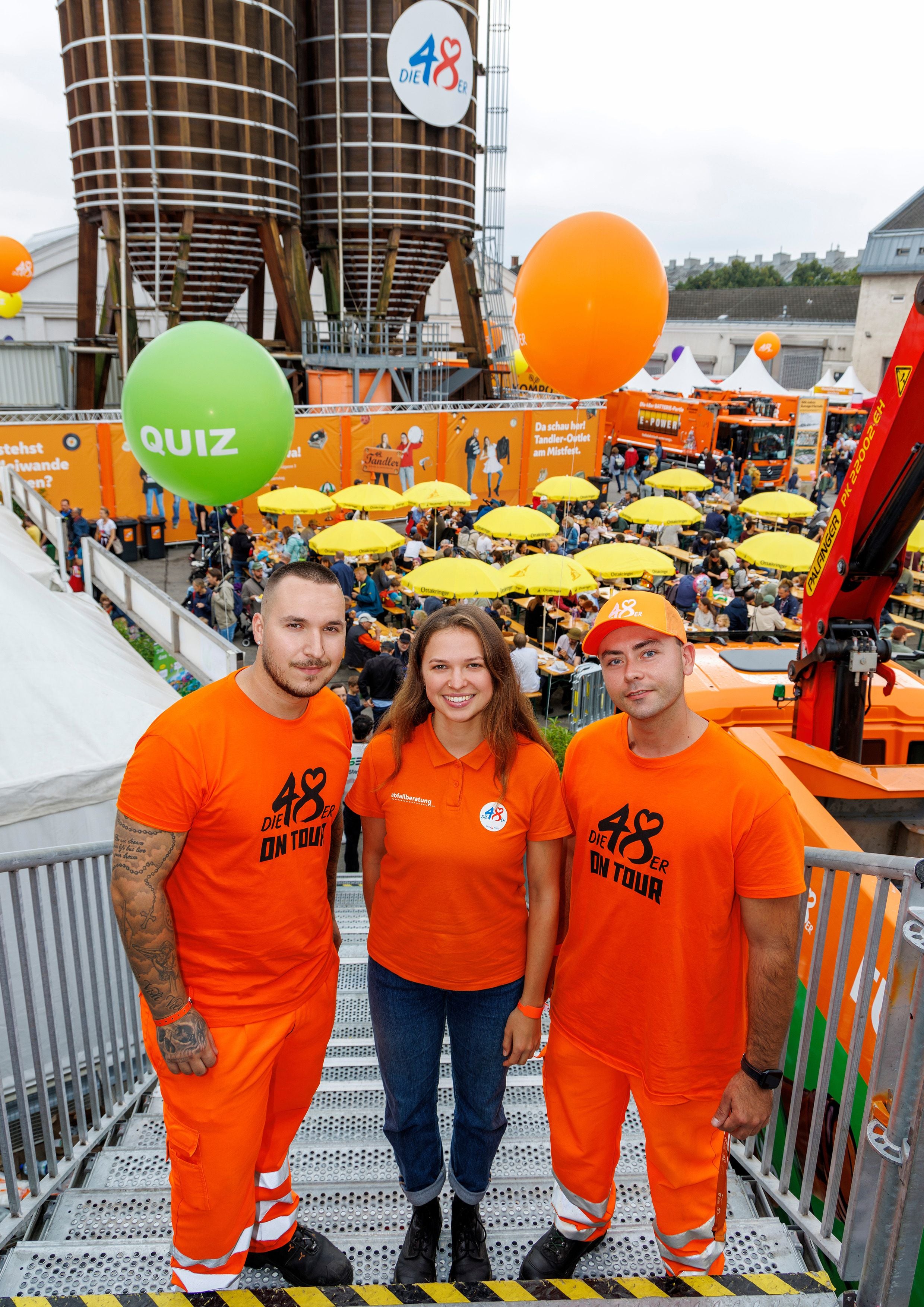Drei Personen in oranger Dienstkleidung posieren beim Mistfest der 48er vor einer Menschenmenge.