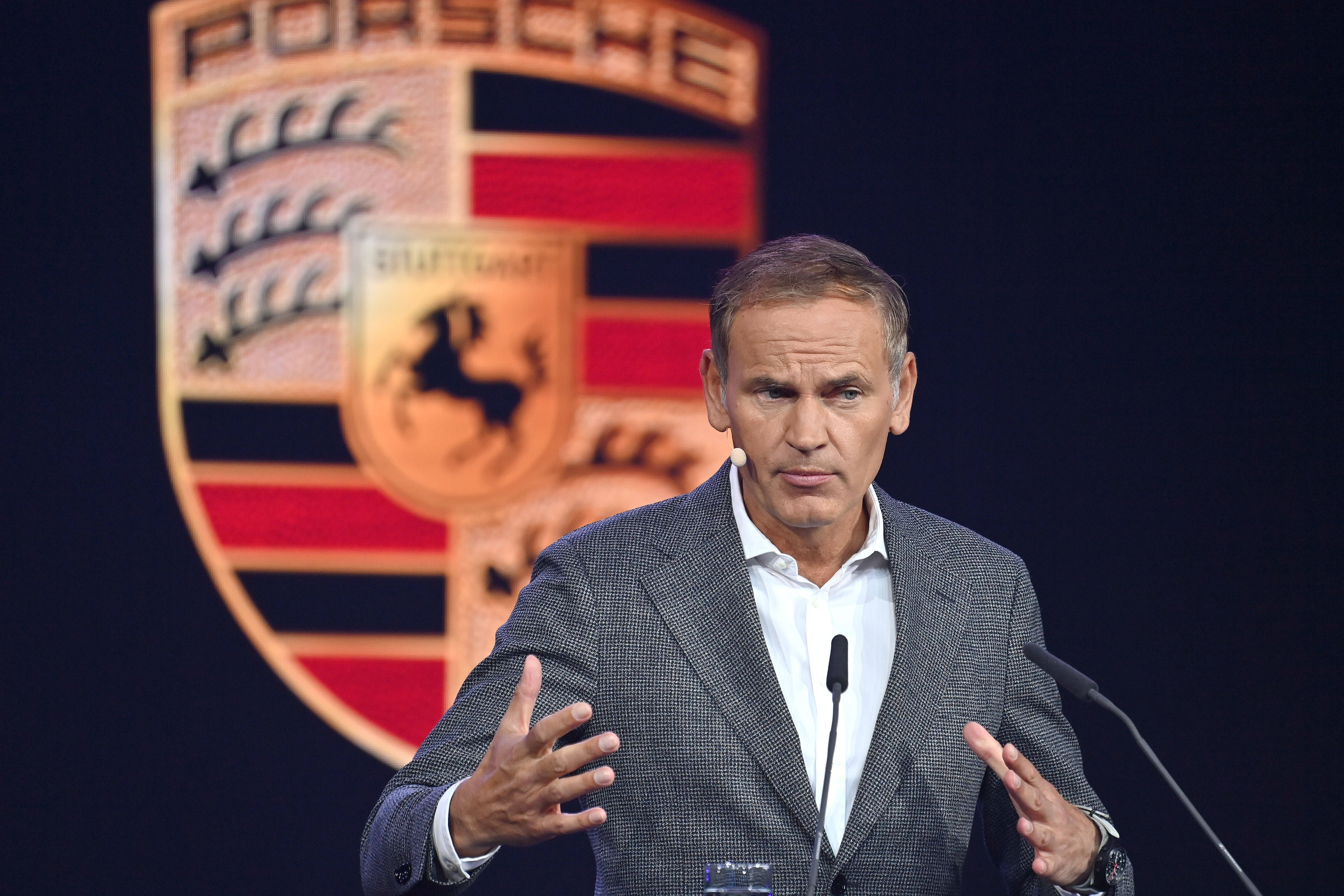 Ein Mann spricht vor Mikrofonen, im Hintergrund ist ein großes Porsche-Logo zu sehen.