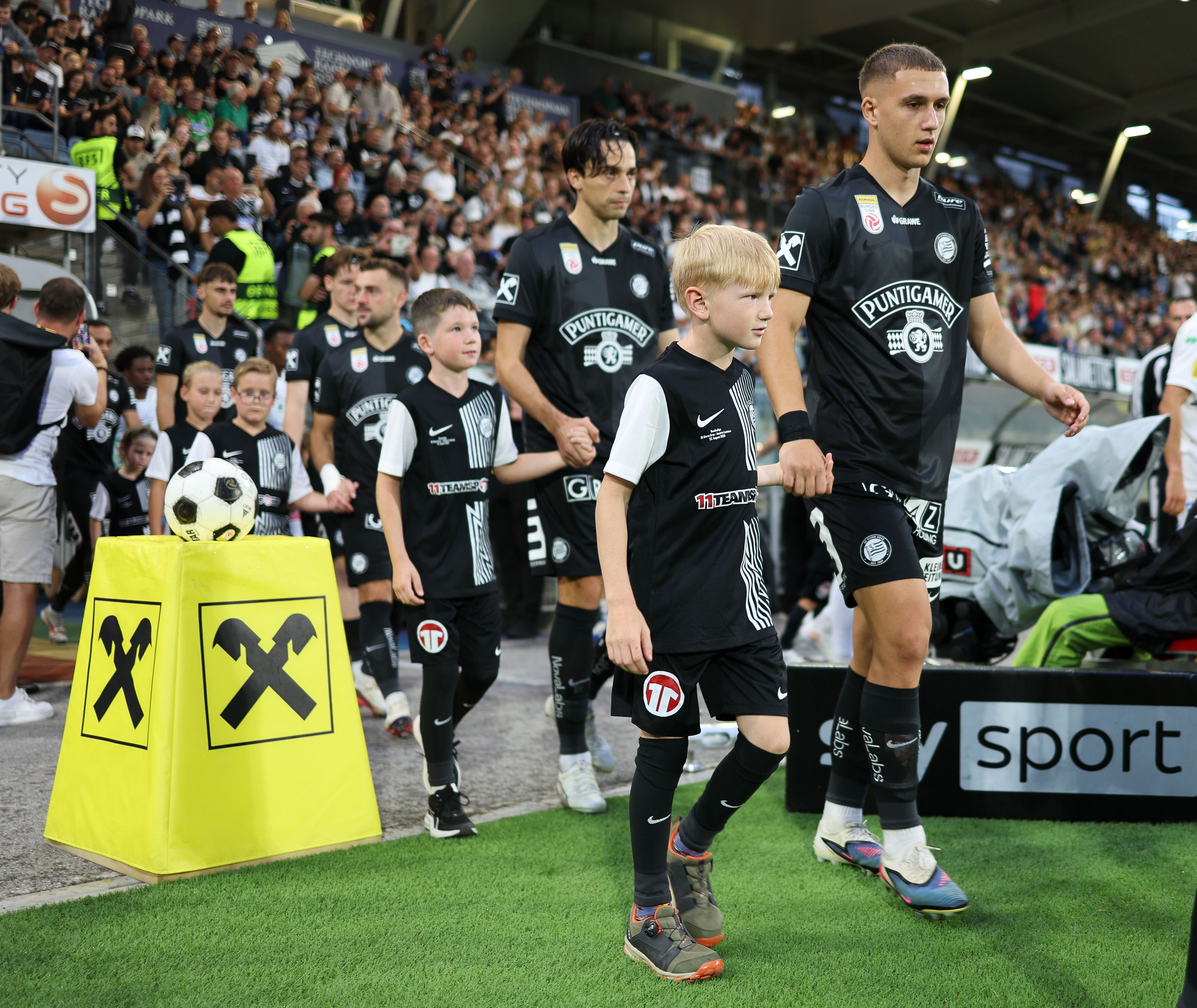 Fußballspieler und Kinder laufen beim Einlaufen ins Stadion, umgeben von vielen Zuschauern.