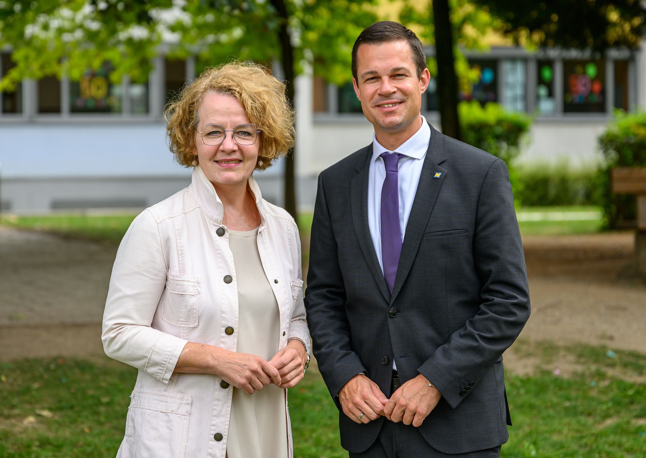 Eine Frau und ein Mann stehen lächelnd im Freien vor einem Gebäude mit Fenstern.