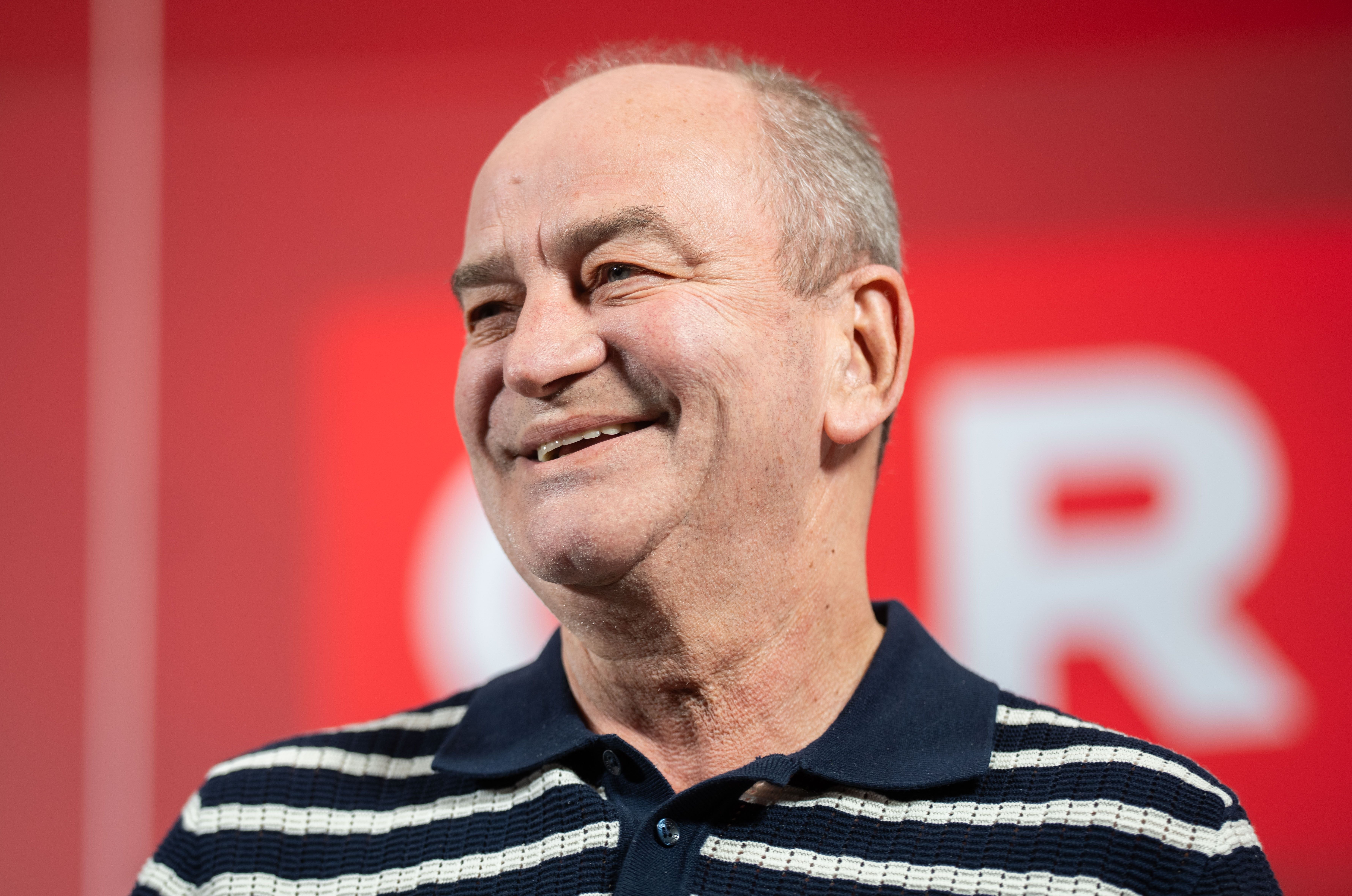 Ein älterer Mann mit gestreiftem Poloshirt lächelt vor rotem Hintergrund.