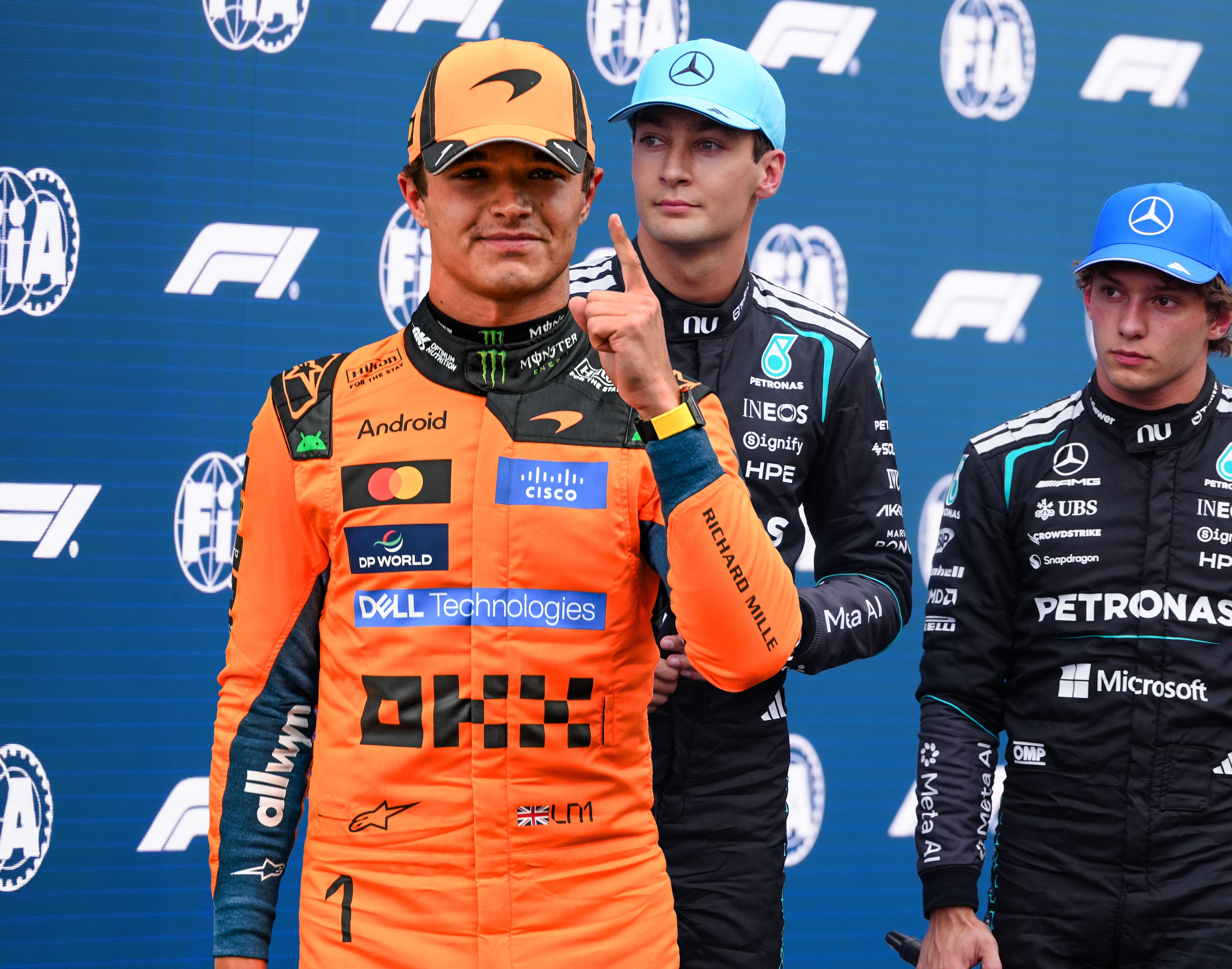 Drei Formel-1-Fahrer posieren vor einer F1-Fotowand, einer zeigt den Finger nach oben.