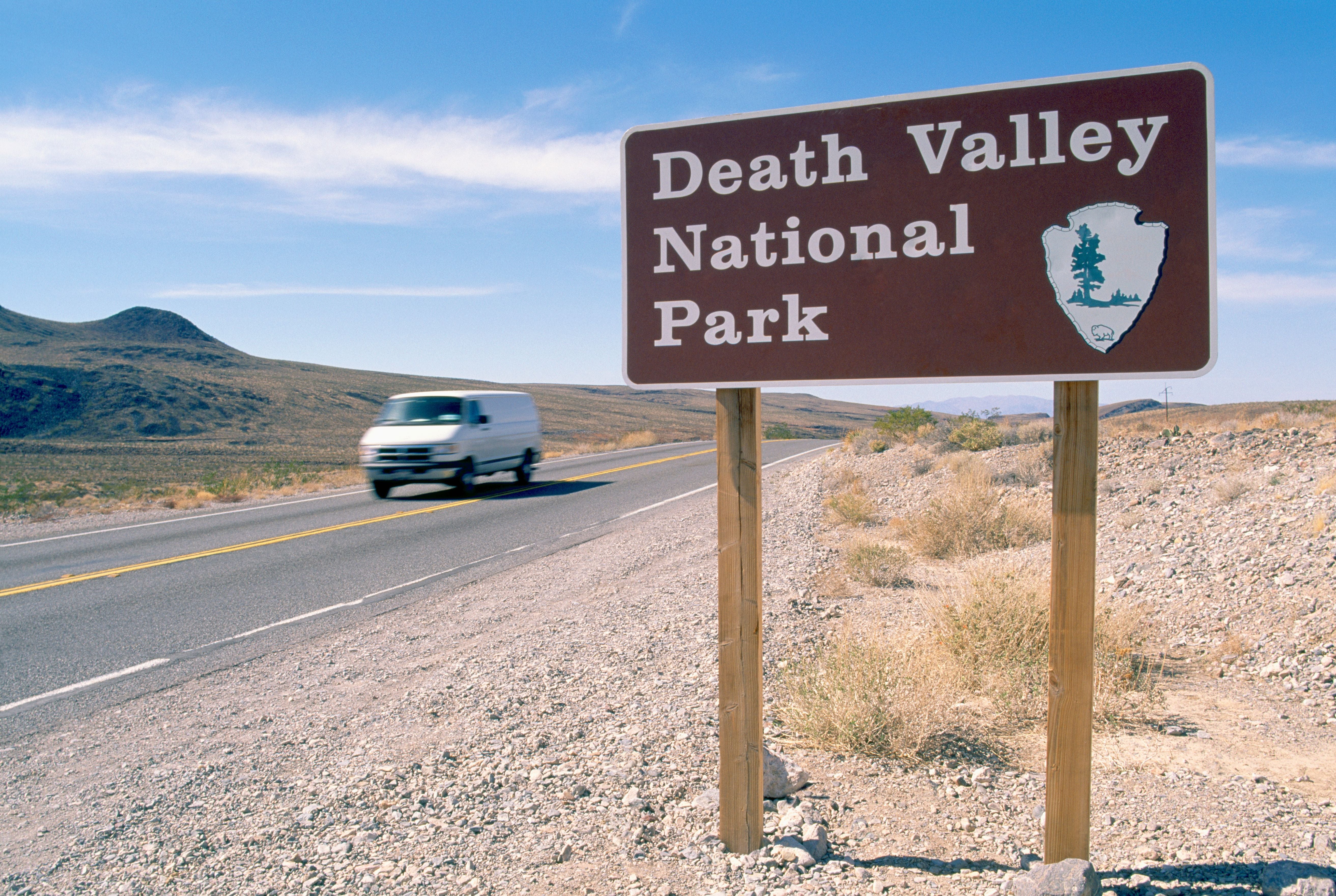 Braunes Schild mit Aufschrift "Death Valley National Park" neben einer Straße in wüstenähnlicher Landschaft.