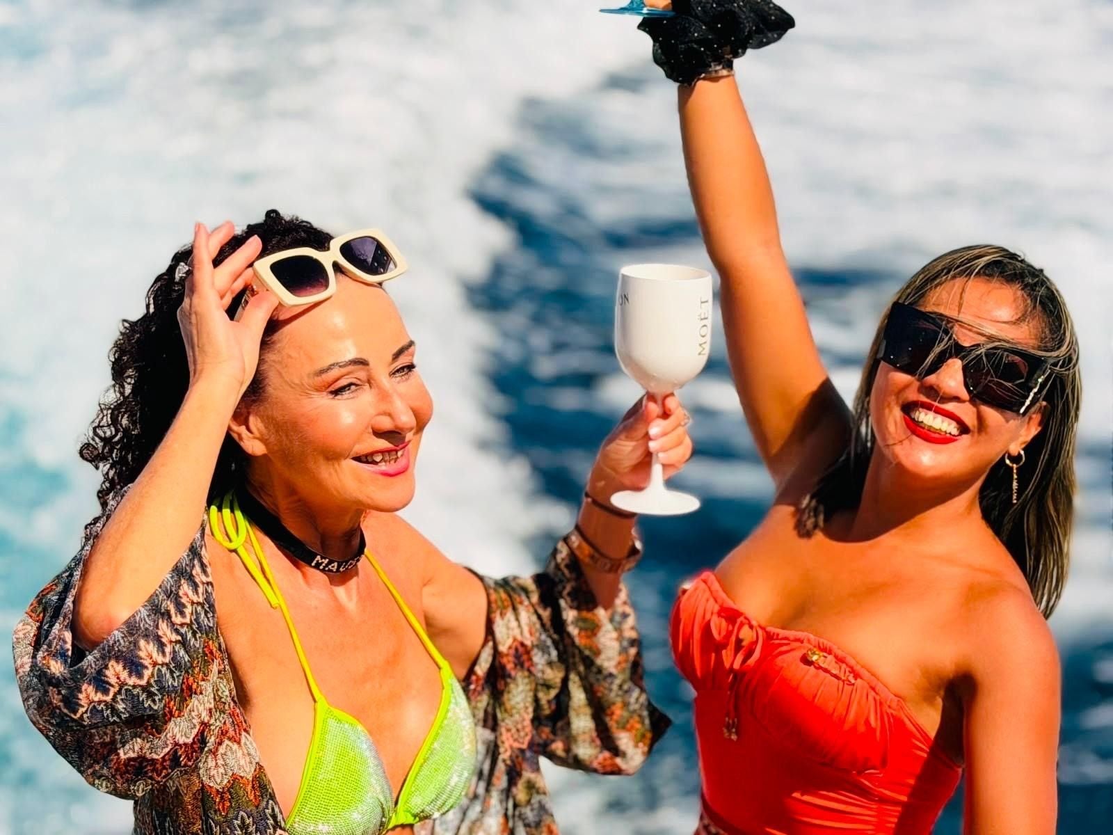 Zwei Frauen feiern lachend auf einer Yacht, eine hält ein Weinglas in der Hand.