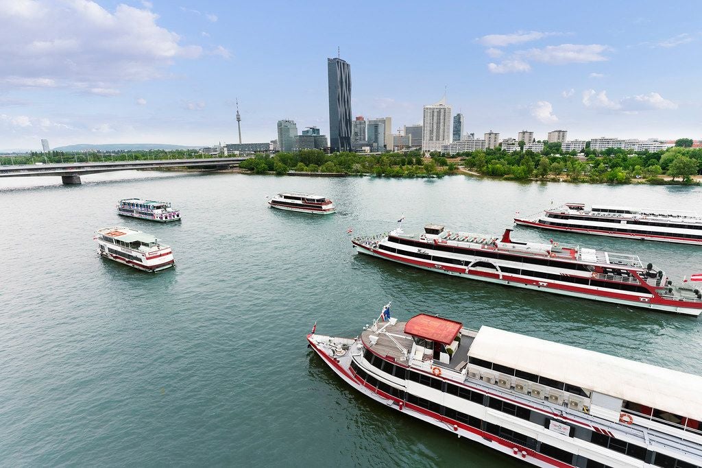Mehrere Ausflugsschiffe fahren auf der Donau vor moderner Stadtkulisse und Brücke.