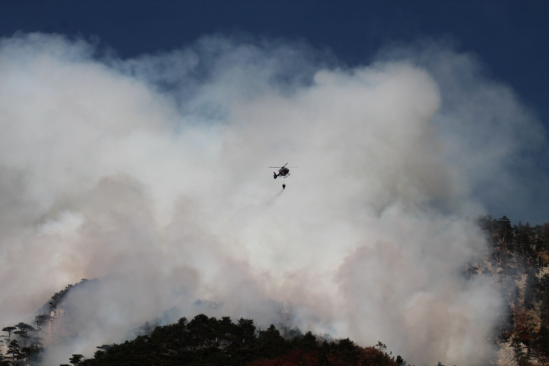 Ein Hubschrauber löscht einen Waldbrand, dichter Rauch steigt über dem Wald auf.